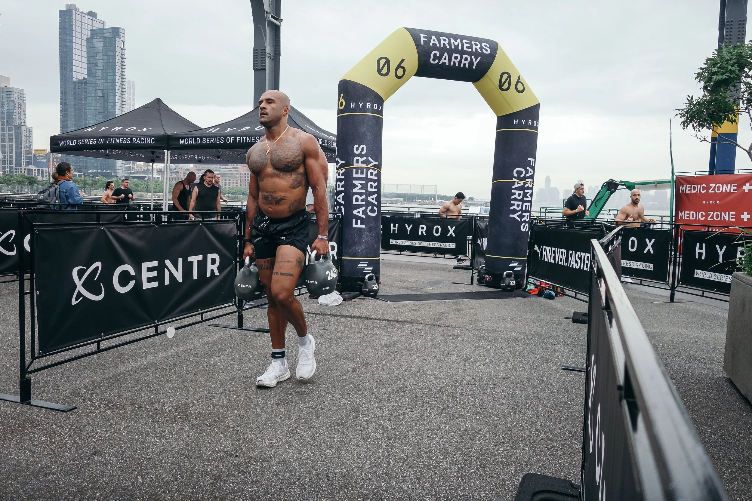A shirtless man with tattoos walking through a fitness competition area, holding kettlebells, with a cityscape background and various banners and tents for the event.