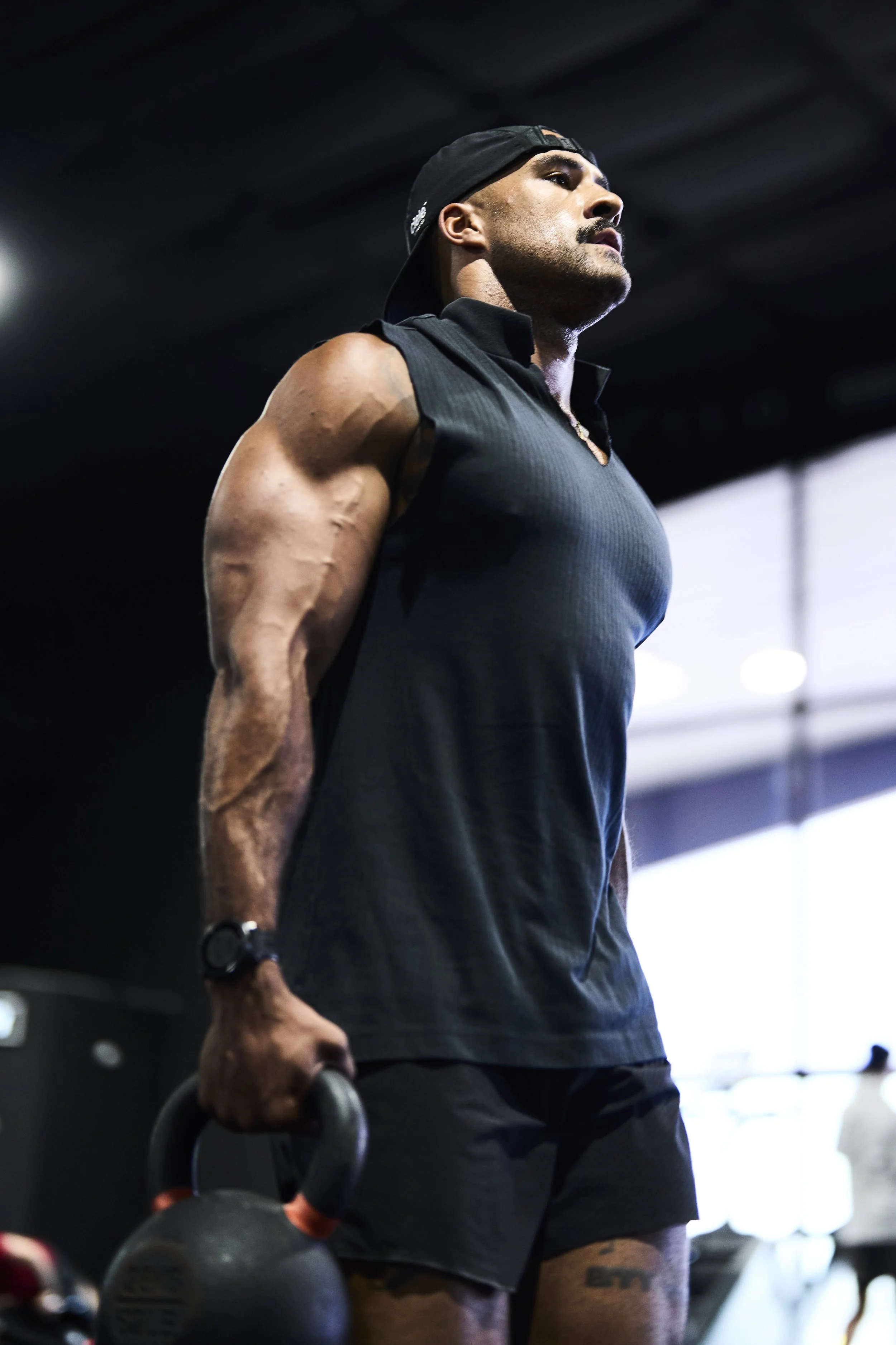 A muscular man in workout attire, holding a kettlebell in one hand, wearing a black tank top, shorts, a cap backward, and a watch, standing in a gym.