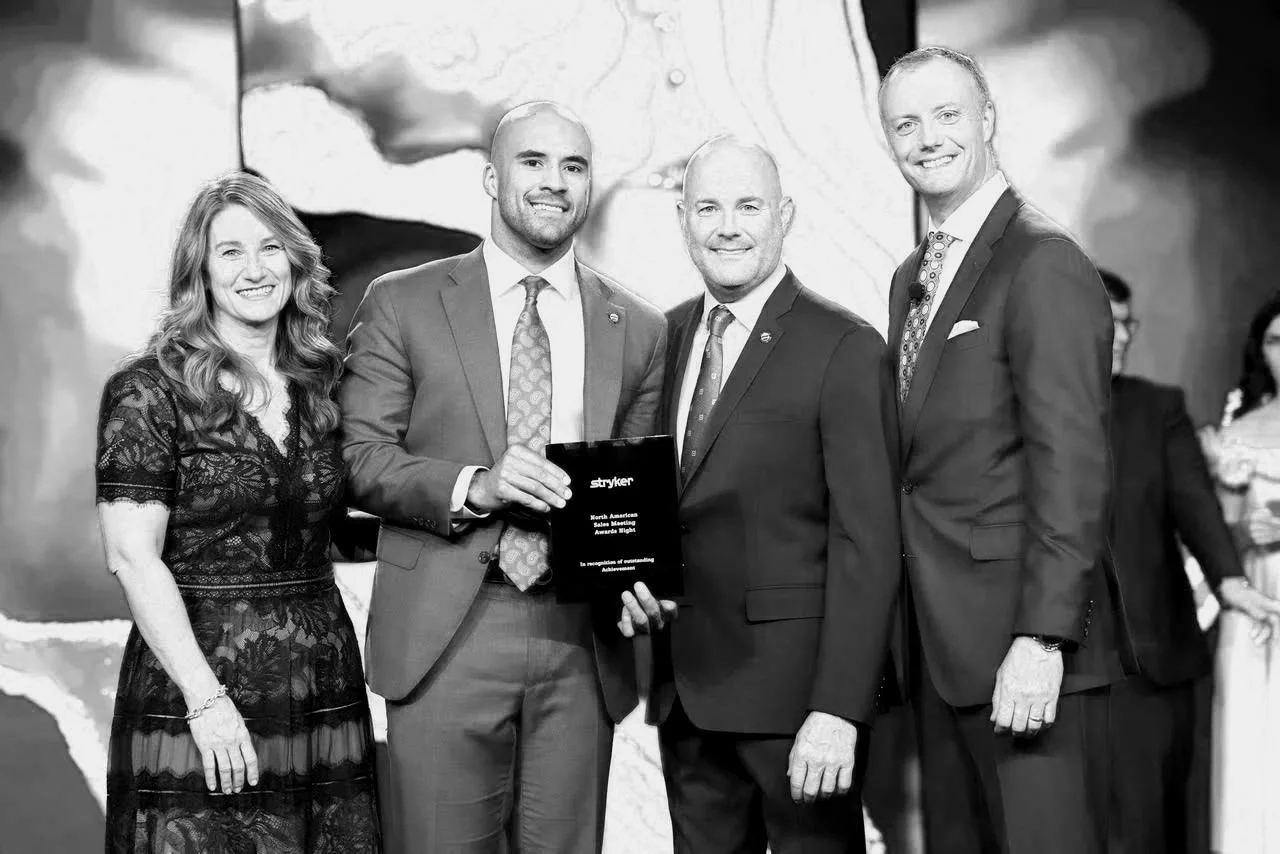 Four people in suits and a woman in a lace dress standing together at an award ceremony, with a man holding an award plaque.