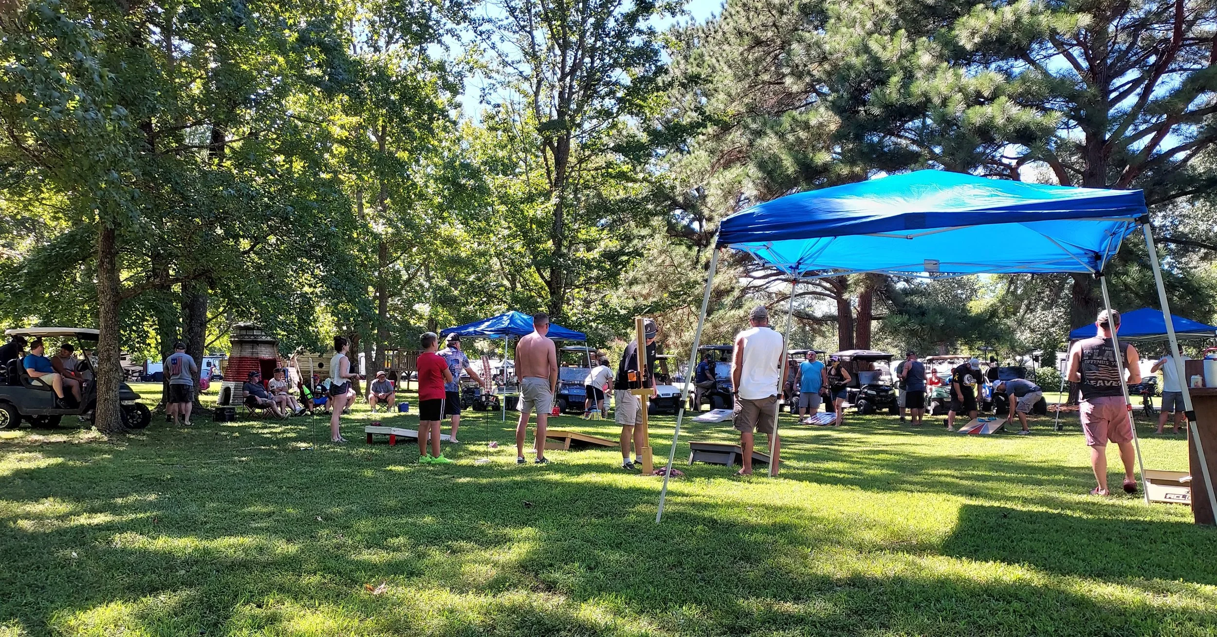 People gathered outdoors in a park with trees, some standing and others sitting, under blue canopies. Golf carts are parked nearby, and various activities and stalls are set up on the grass.
