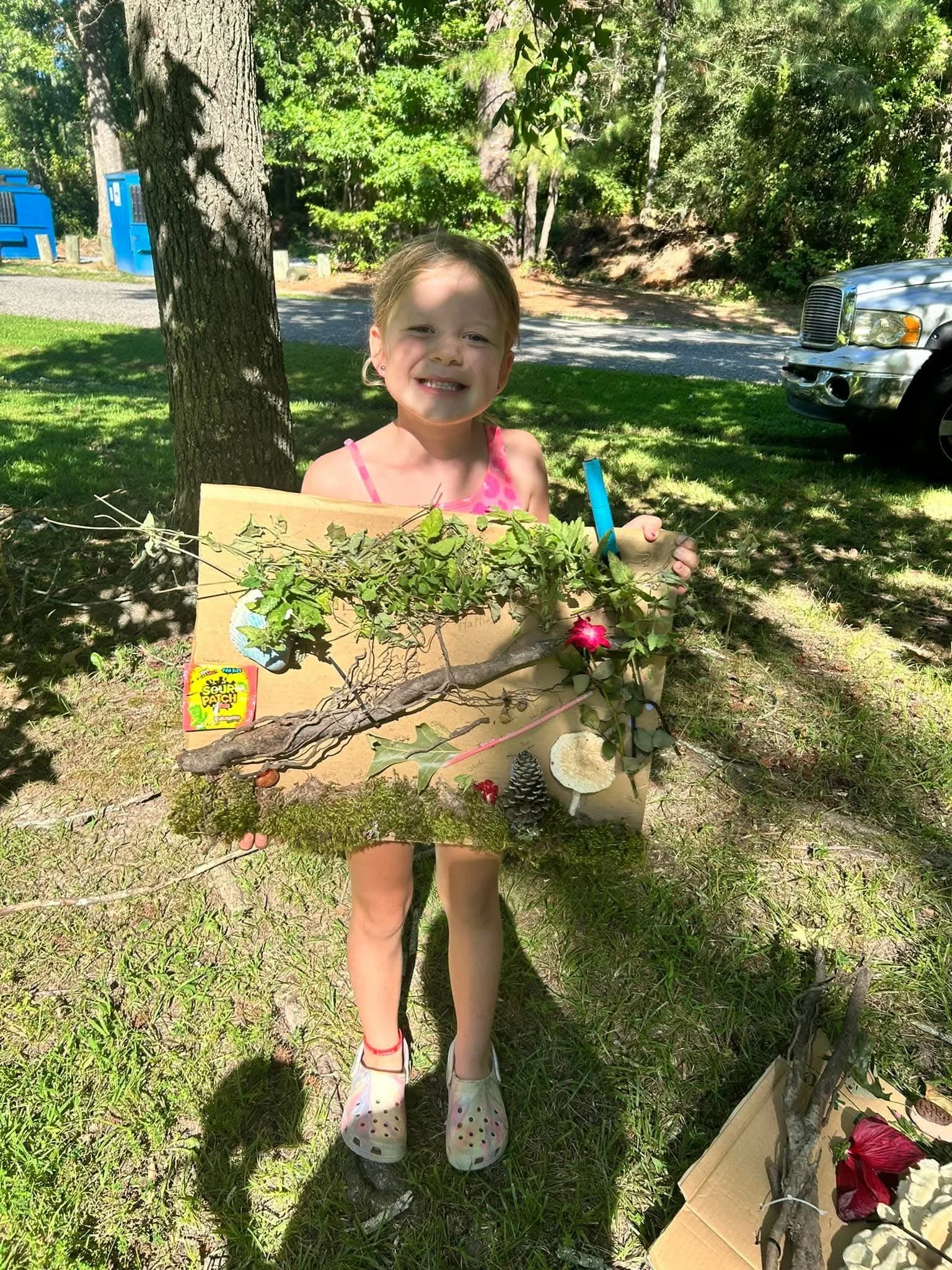 A young girl with a big smile holding a decorated artistic display of tree branches, leaves, pinecones, and a pink flower, outdoors on a sunny day with trees, grass, and a parked vehicle in the background.