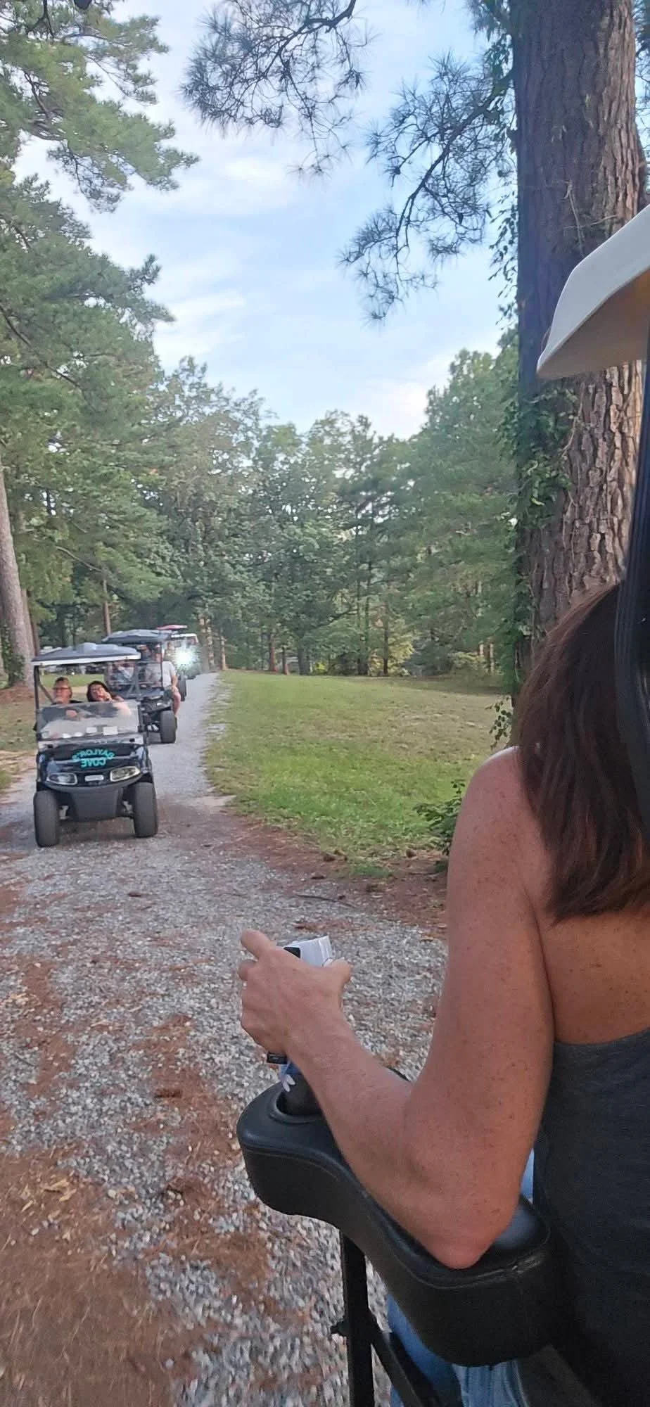 Line of golf carts on a gravel trail through a wooded area, with one person steering the cart on the right side of the image.