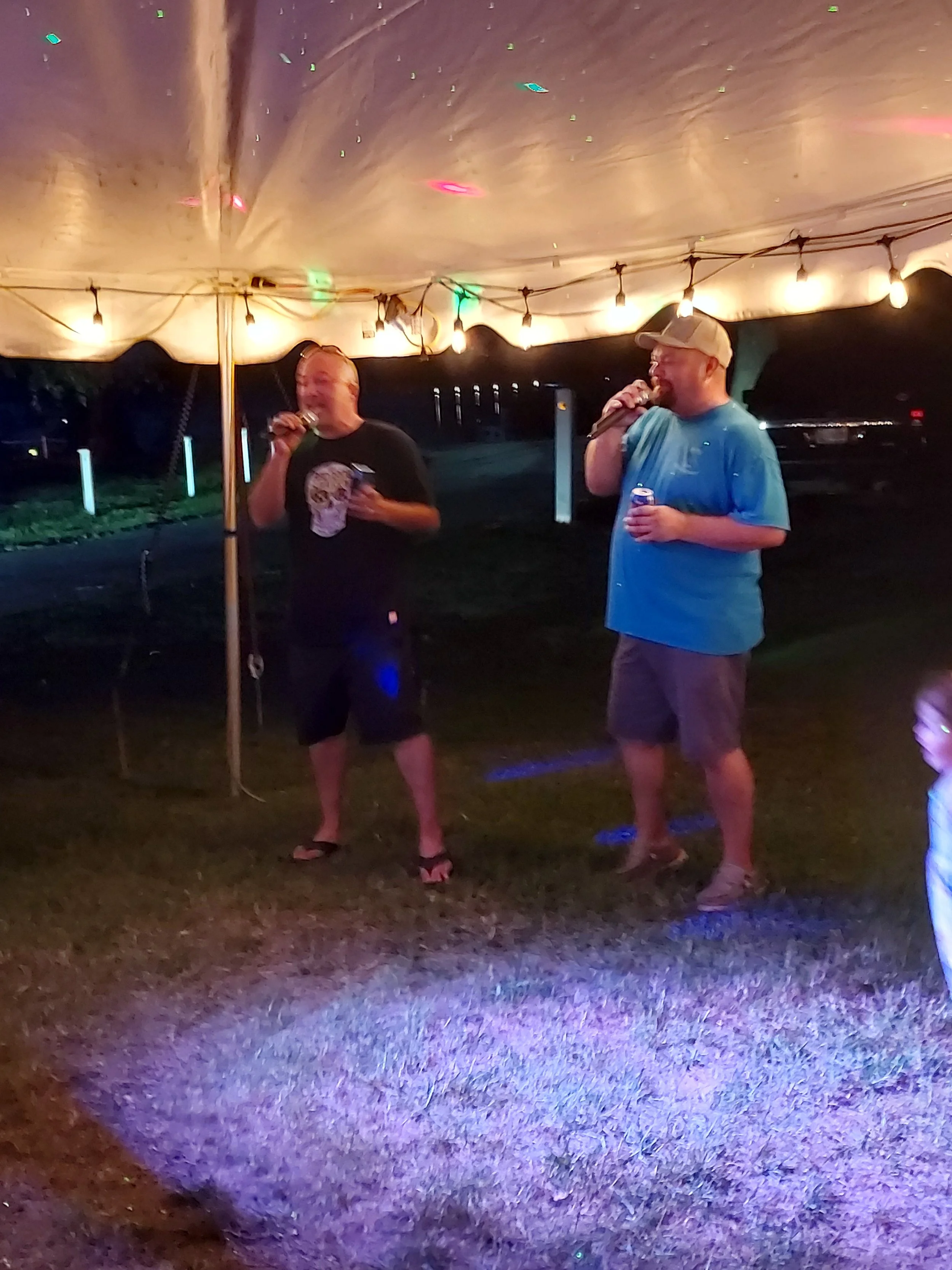 Two men standing under a string of lights at night, both holding microphones and drinks, in a casual outdoor setting.