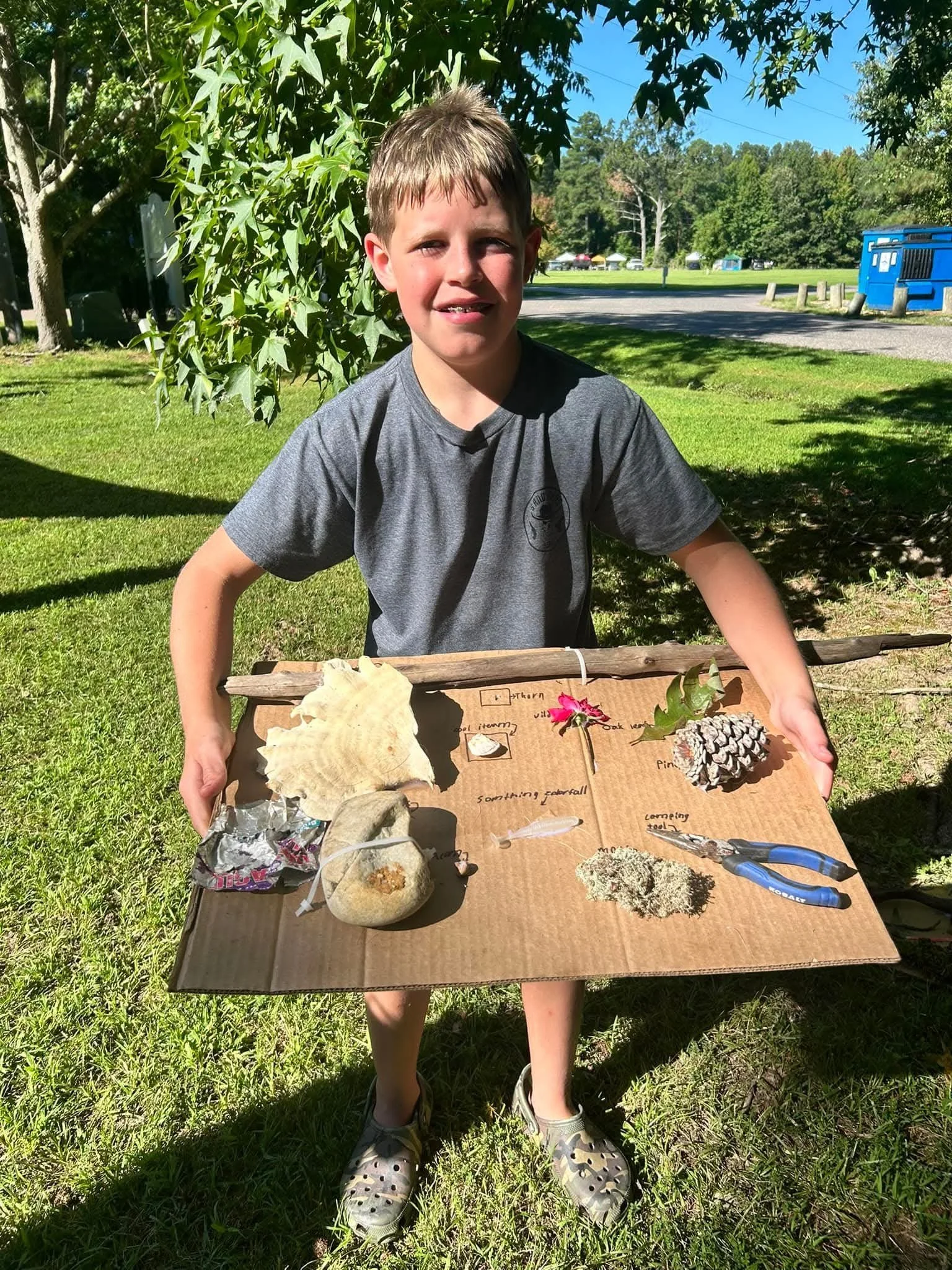 A young boy with short light brown hair and wearing a gray T-shirt and Crocs shoes is standing outdoors on grass, holding a cardboard display board with various natural objects attached to it. The background features trees, a parking lot, and a blue 