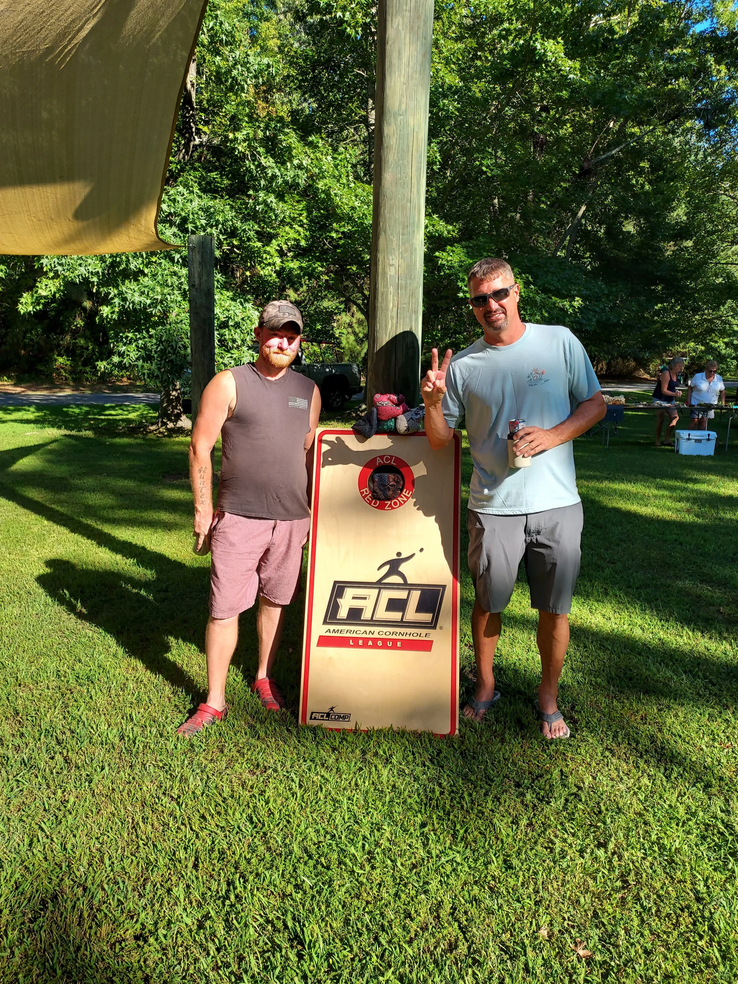 Two men standing outdoors in front of a sign that reads 'ACL - American Cornhole League.' One man is wearing sunglasses, a light blue T-shirt, and shorts, making a peace sign with his right hand. The other man is wearing a sleeveless shirt, shorts, a
