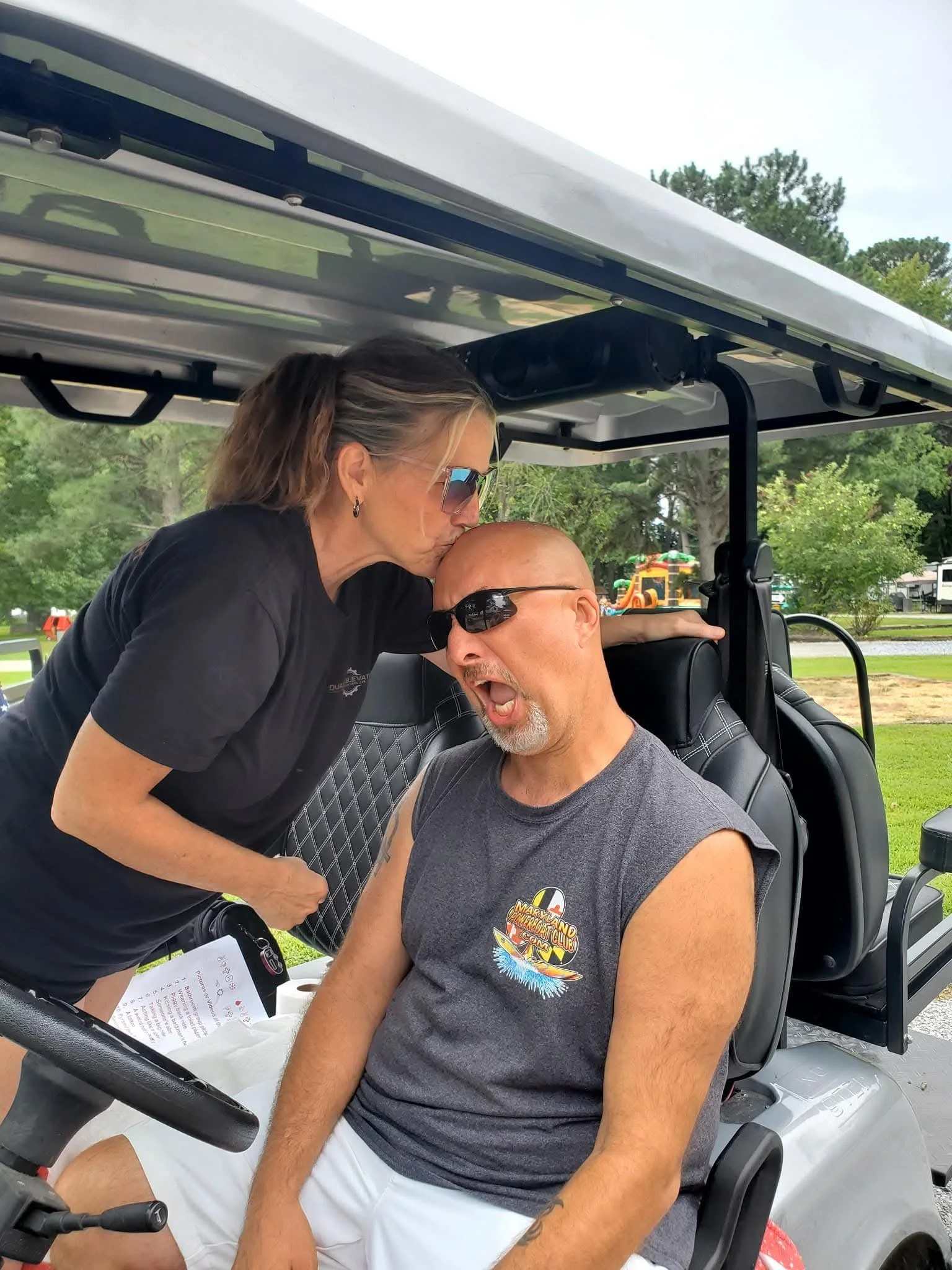 A woman kissing a man on the head while sitting in a golf cart outdoors. The man has his eyes closed and is wearing glasses and a sleeveless shirt. The woman is wearing sunglasses and earrings. There is a grassy area, trees, and some playground equip