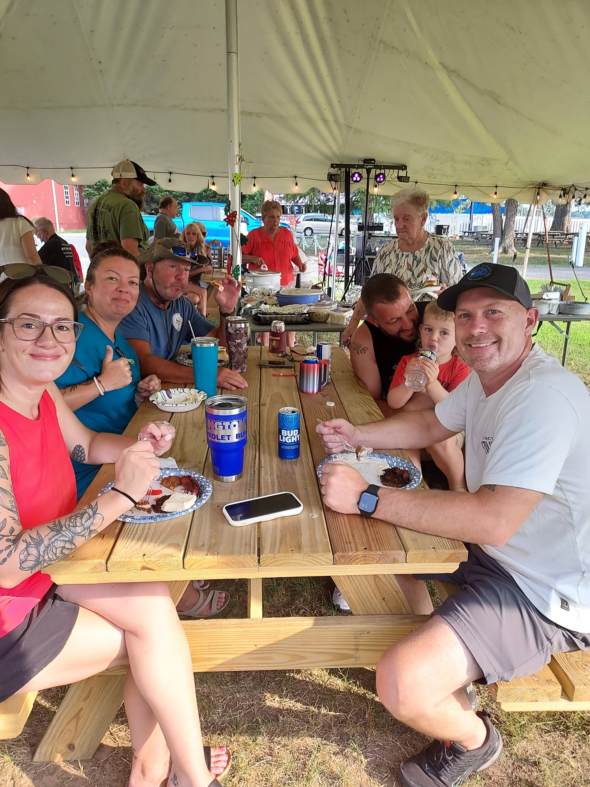 Gathering of people at a picnic table under a tent, enjoying food and drinks, with some looking at the camera and others engaged in conversation, at an outdoor event.