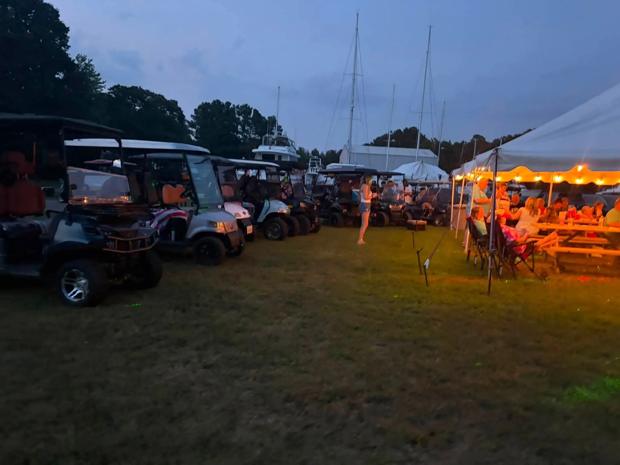 A scene at a marina with several boats docked, some outdoor seating with string lights, and a group of people gathered under a tent during evening or dusk.
