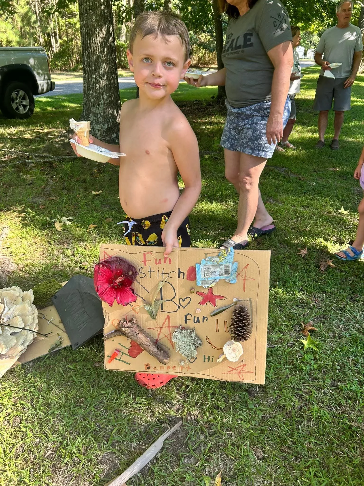 A young boy holding a plate with an ice cream cone, standing outdoors on grass next to a colorful handmade poster decorated with various natural objects like flowers, pinecone, and rocks, at a park or picnic area with other people in the background.