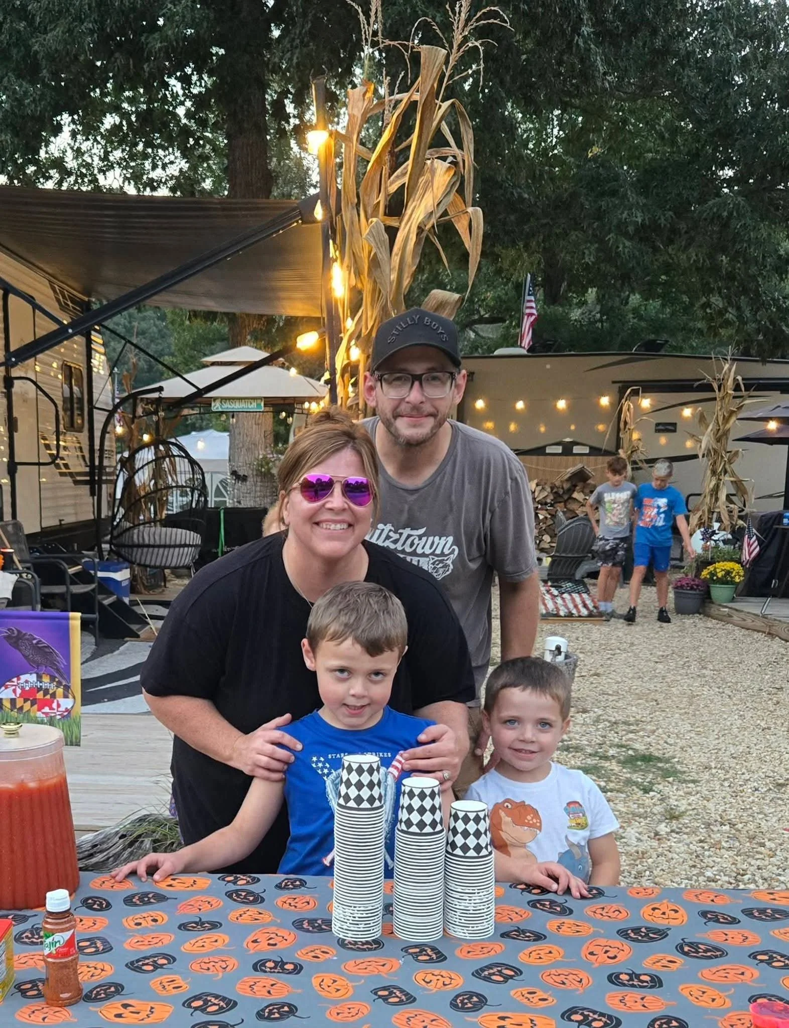 A group of people, including children, at a Halloween-themed outdoor gathering. The table in front has a Halloween themed tablecloth, and there are cups and a bottle of sauce on it. Behind them, there are autumn decorations with corn stalks and light