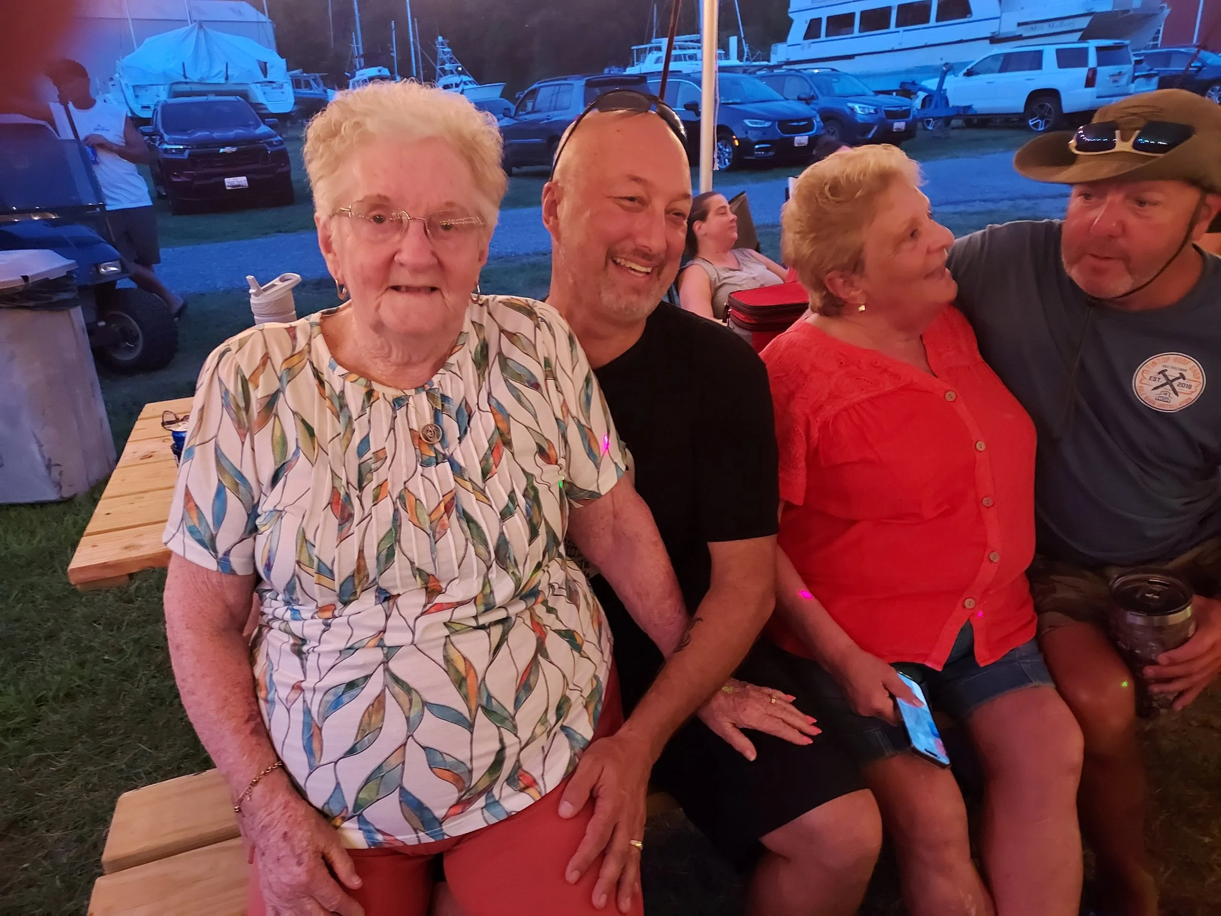 People sitting on benches at an outdoor gathering near a marina with boats and parked vehicles in the background, during evening or night.
