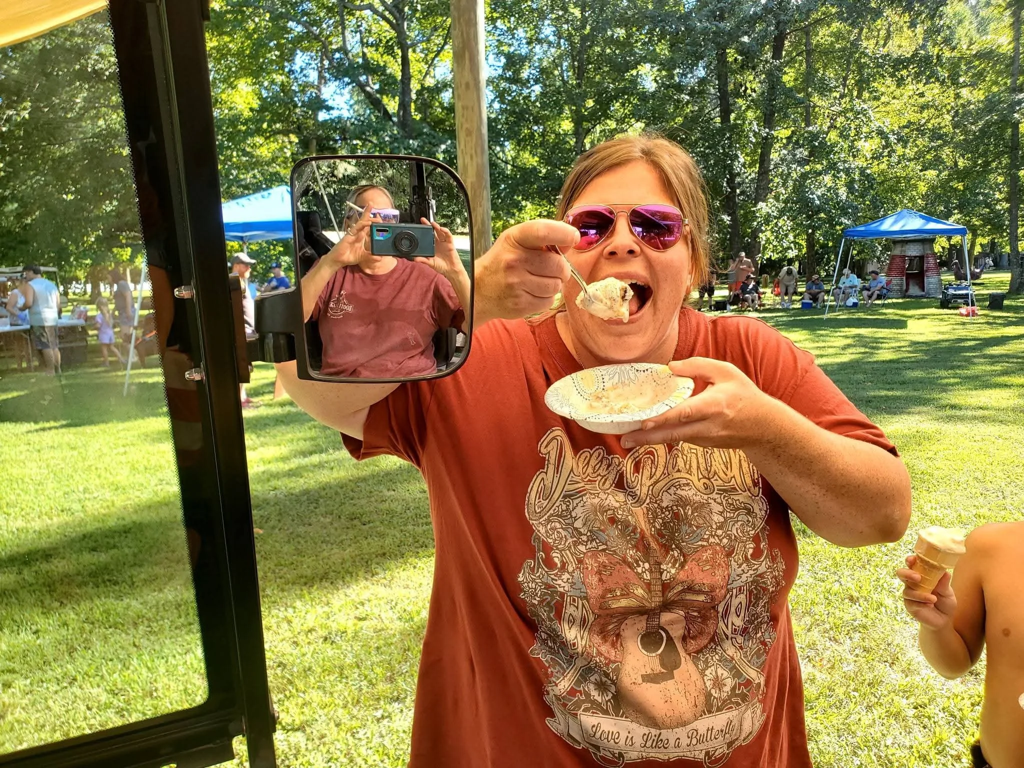 A woman with short hair wearing pink sunglasses is eating ice cream at an outdoor event. Another person, visible in the mirror of a vehicle, is taking a photo of her with a camera. There are trees, a blue canopy, and people in the background.
