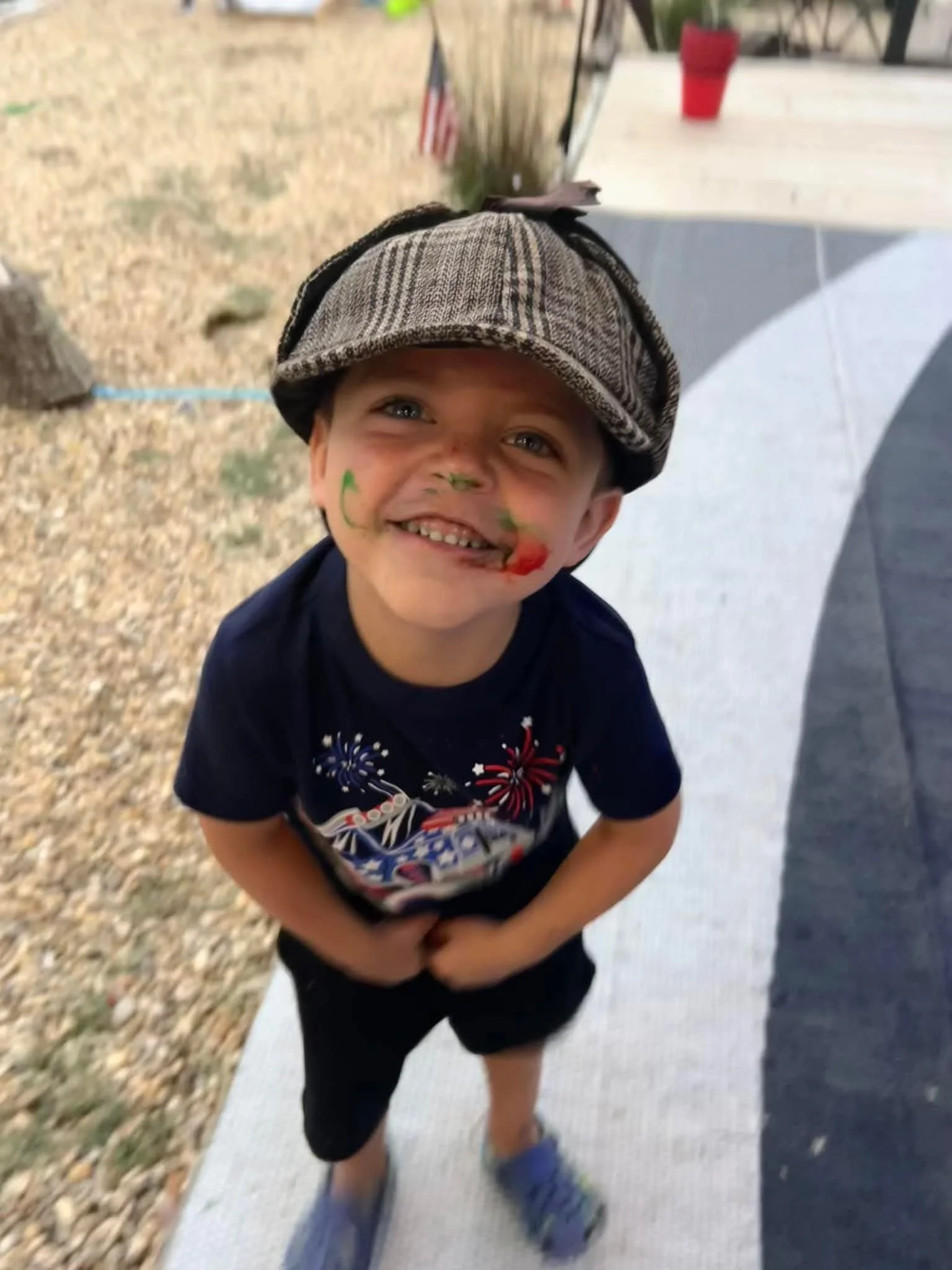 Young boy smiling with face paint, wearing a hat, and standing on a sidewalk outdoors.
