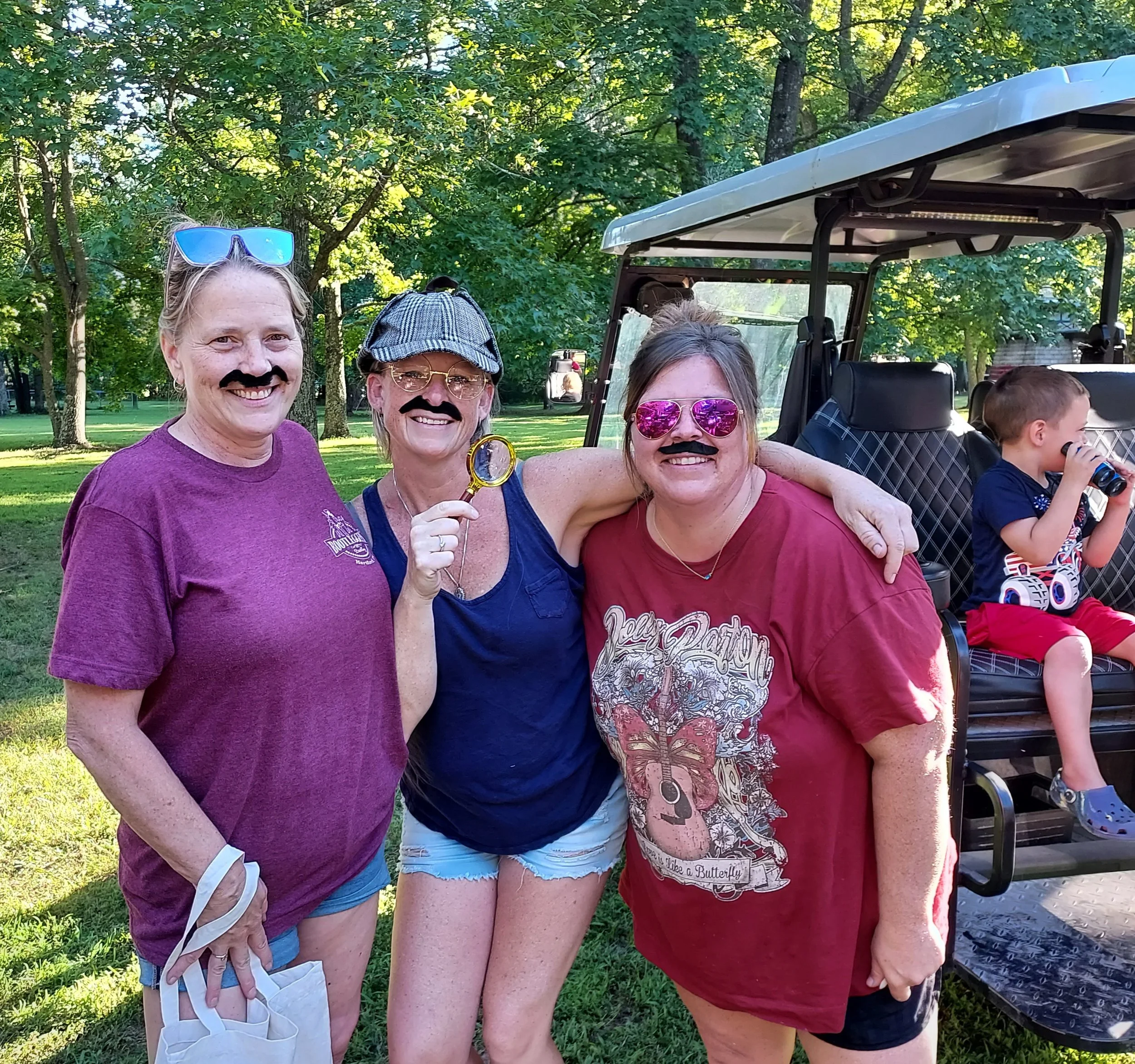 Group of three women with pretend mustaches and sunglasses, smiling outdoors during daytime, with trees in the background and a golf cart nearby; a young boy with a water bottle is partially visible on the right.