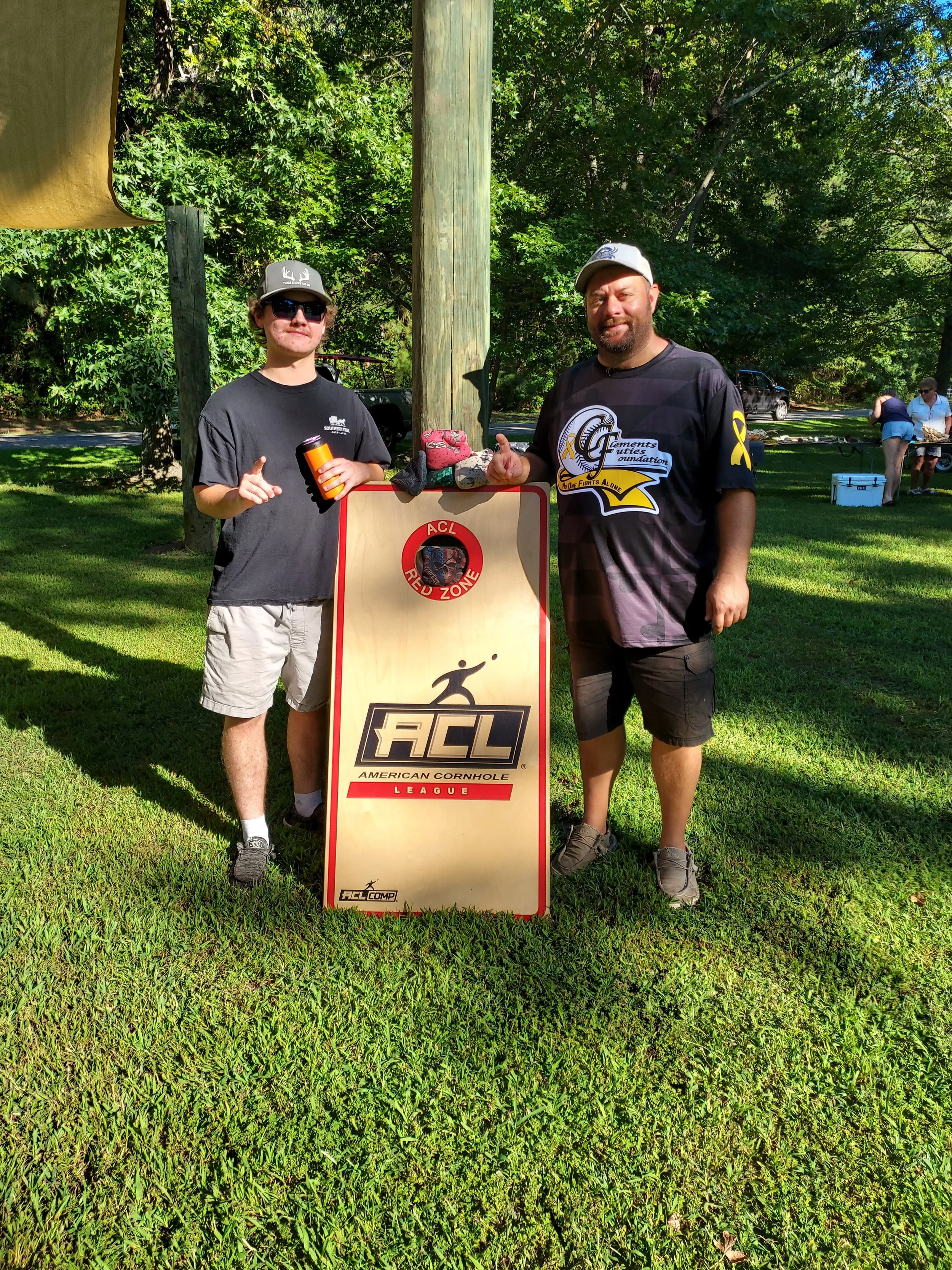 Two men standing outdoors on grass, posing beside a sign that reads 'ACL American Cornhole League.' One man is wearing sunglasses, a cap with a deer antler logo, and a dark shirt, while the other man is wearing a cap, a black shirt with a logo, and s