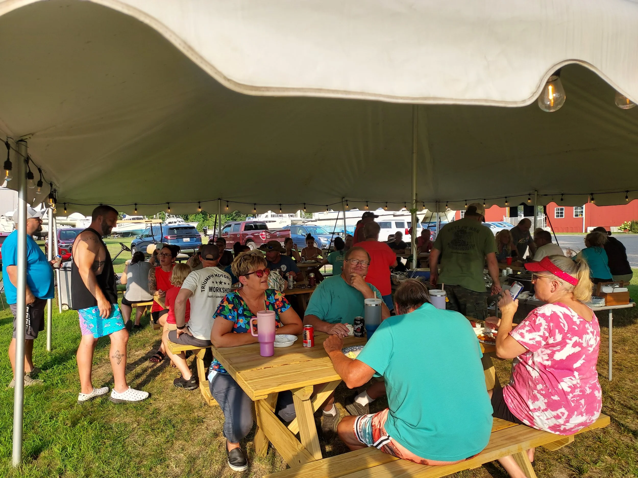People gathered under a large tent, sitting at picnic tables, enjoying food and conversation at an outdoor event, with cars and buildings in the background.