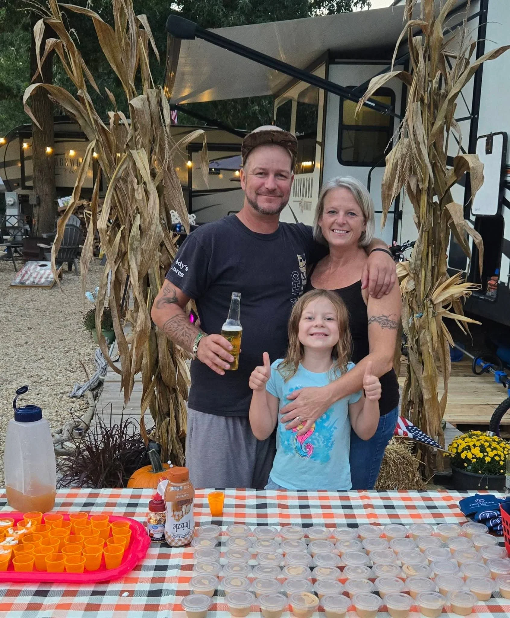 A smiling man, woman, and girl at a fall-themed outdoor event standing behind a table with orange shot glasses and small cups of pudding, surrounded by autumn decorations like dried corn stalks and pumpkins.
