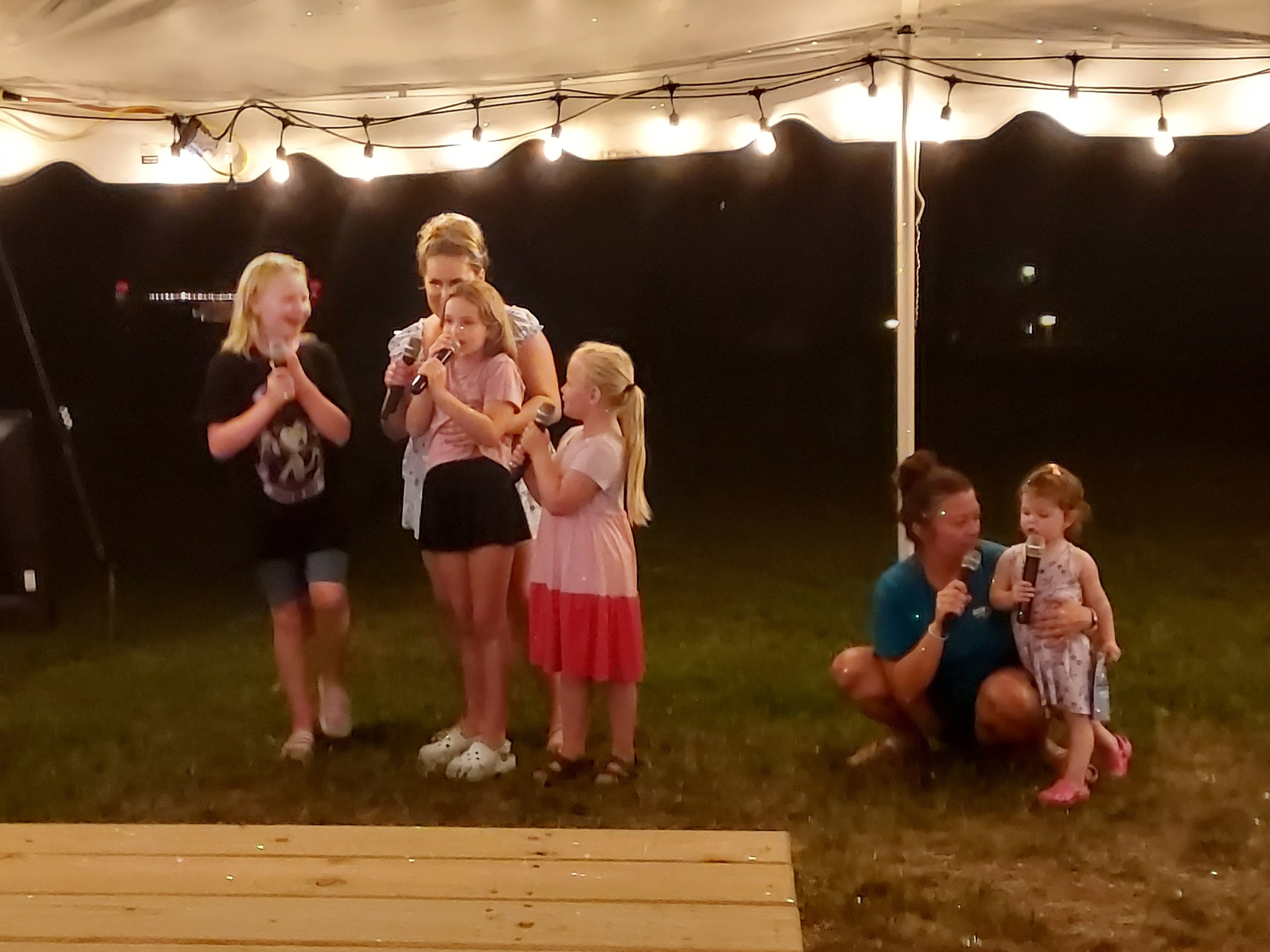 Children and adults at a nighttime outdoor event standing on grass under string lights, some holding microphones, some smiling and interacting.