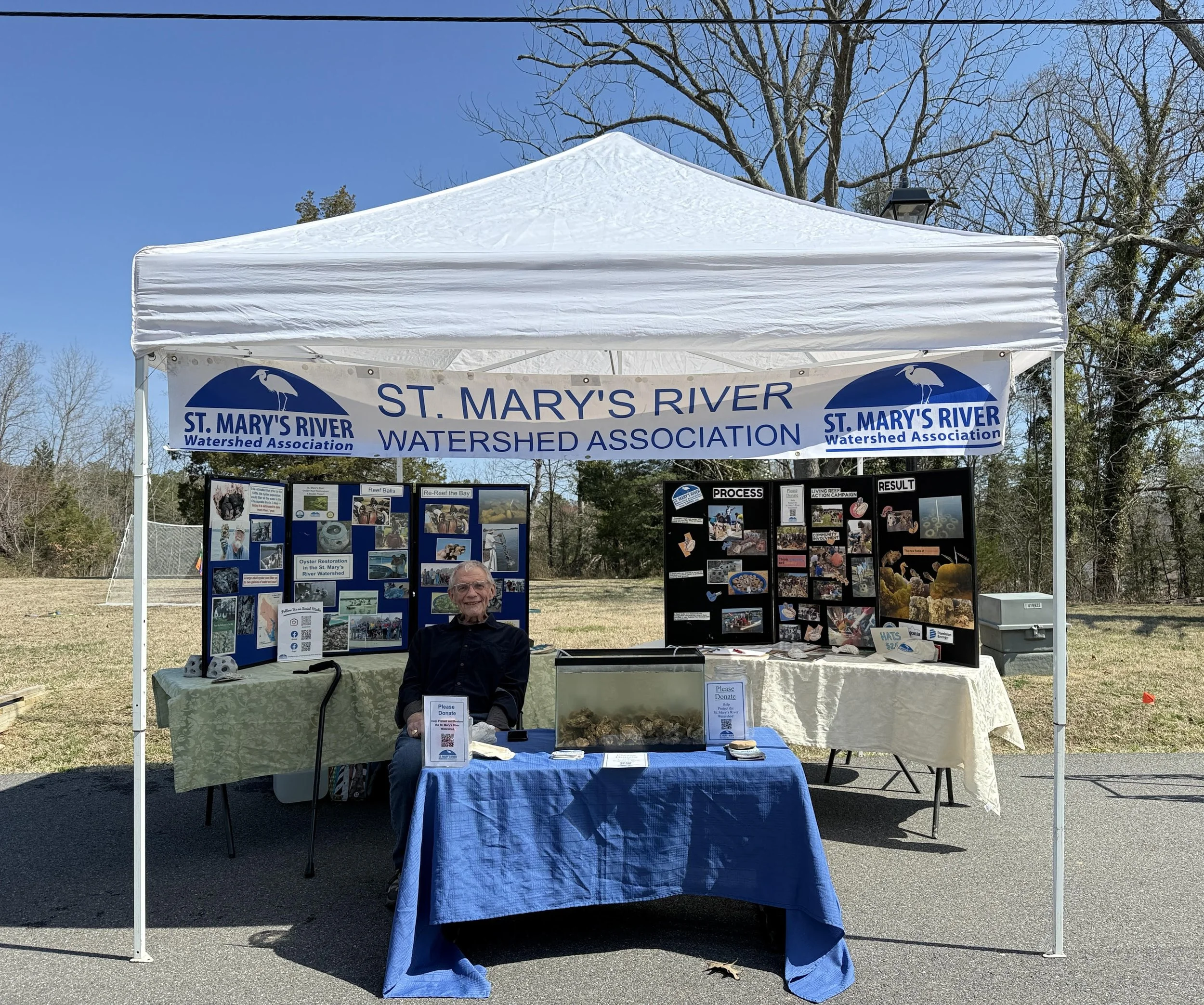 A booth at an outdoor event for St. Mary's River Watershed Association with informational displays and a person sitting at the table.