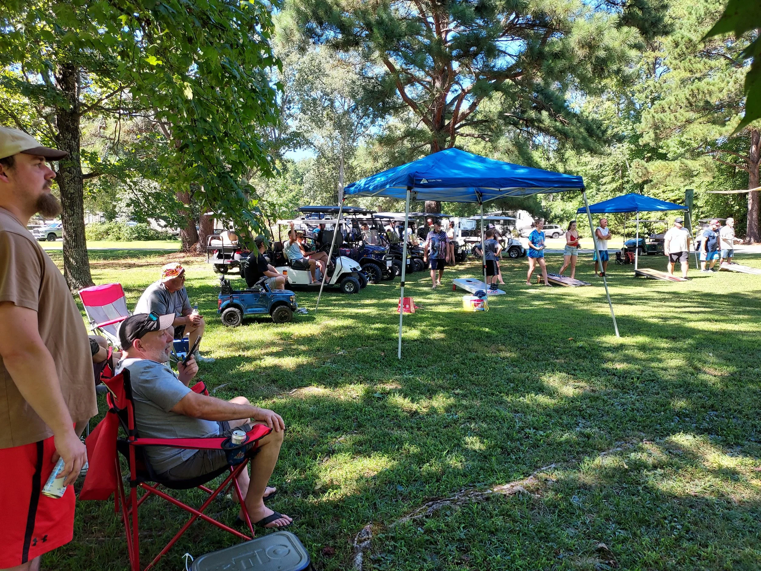 A group of people gathered outdoors in a park-like setting with trees, some seated on foldable chairs and others standing near a blue canopy tent, with golf carts and tents in the background.