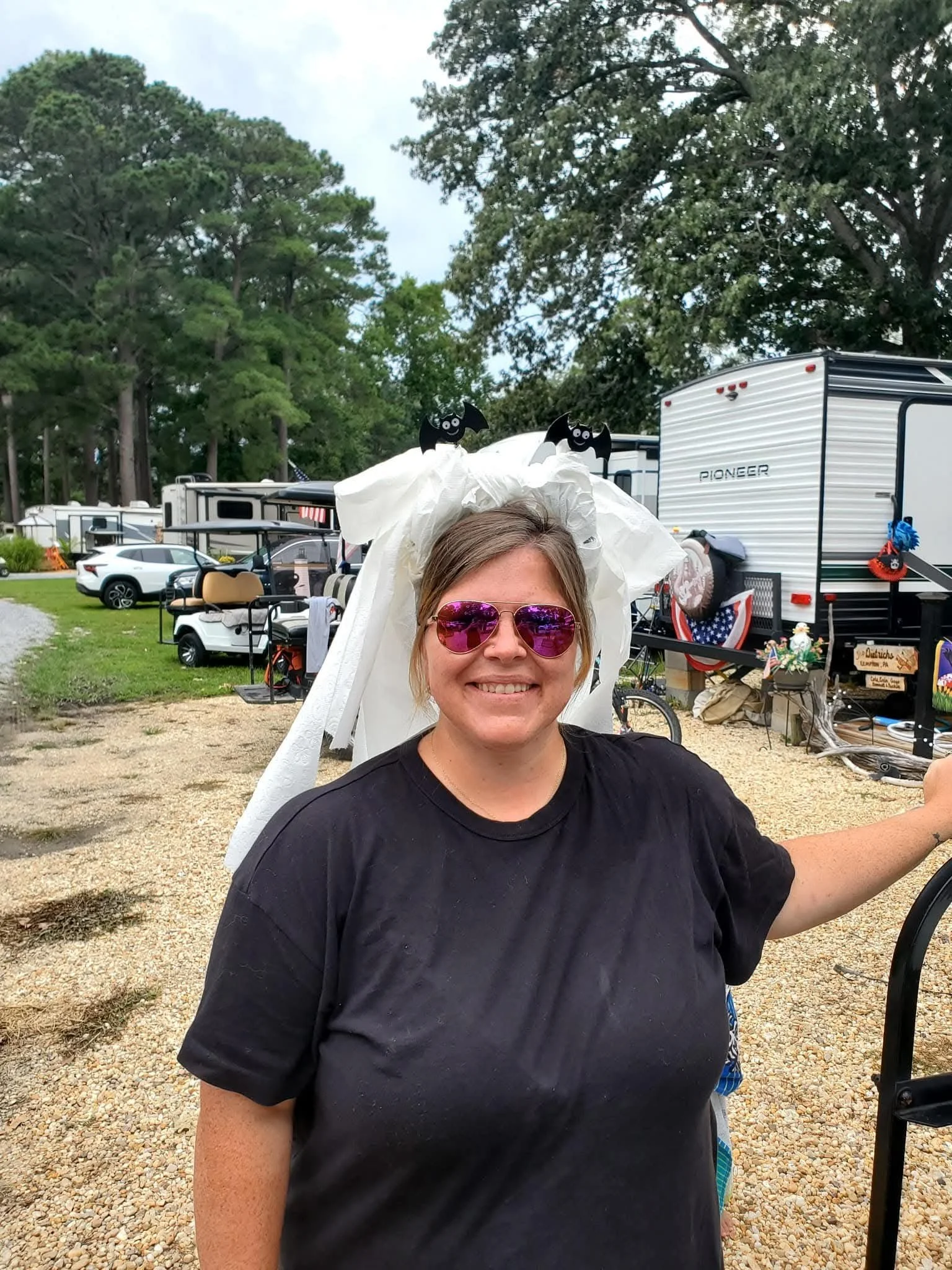 A woman smiling and wearing pink sunglasses, standing outside in a campground area with trees, RVs, and golf carts in the background.