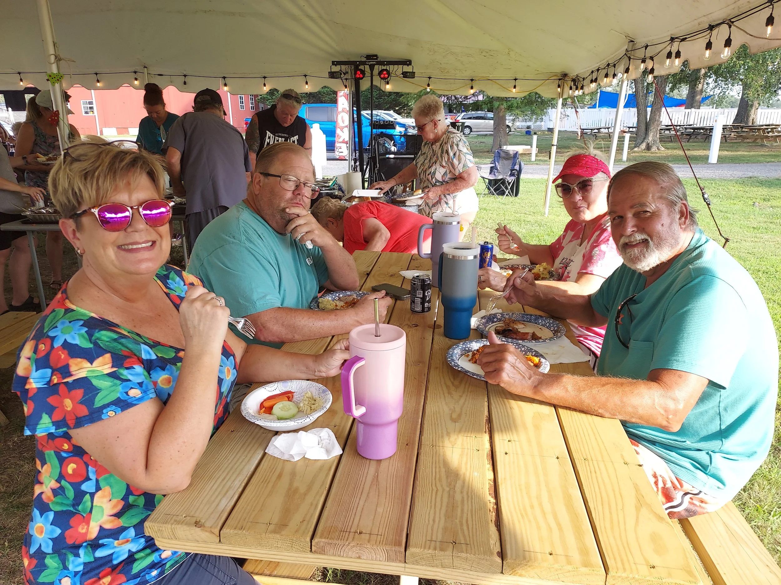 People sitting at a long outdoor table under a tent, enjoying a meal with plates of food, drinks, and cutlery, during a sunny day.