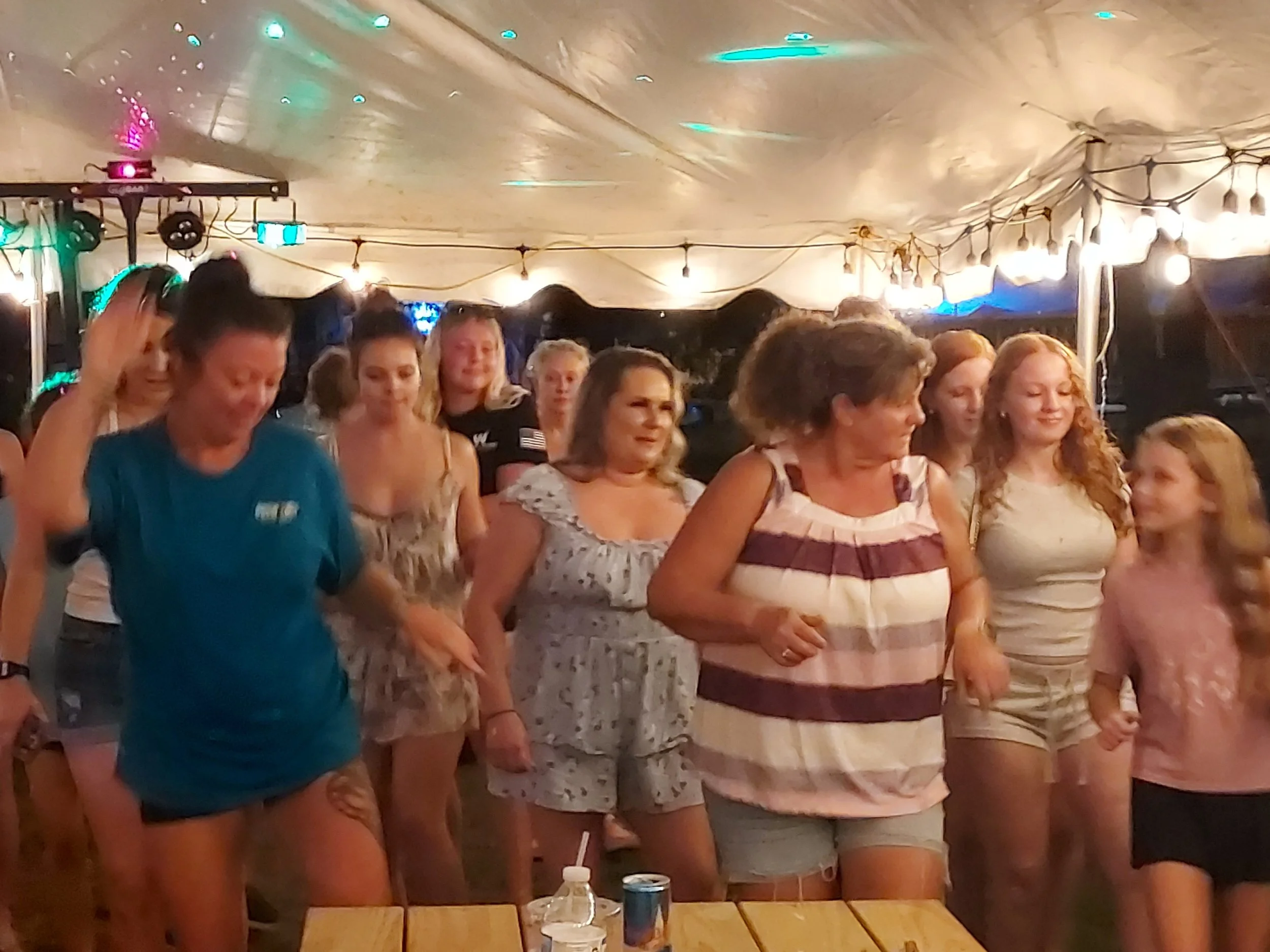 Group of women and girls standing together at an indoor party, some smiling and others looking at something out of frame, with string lights and a tent-like ceiling in the background.