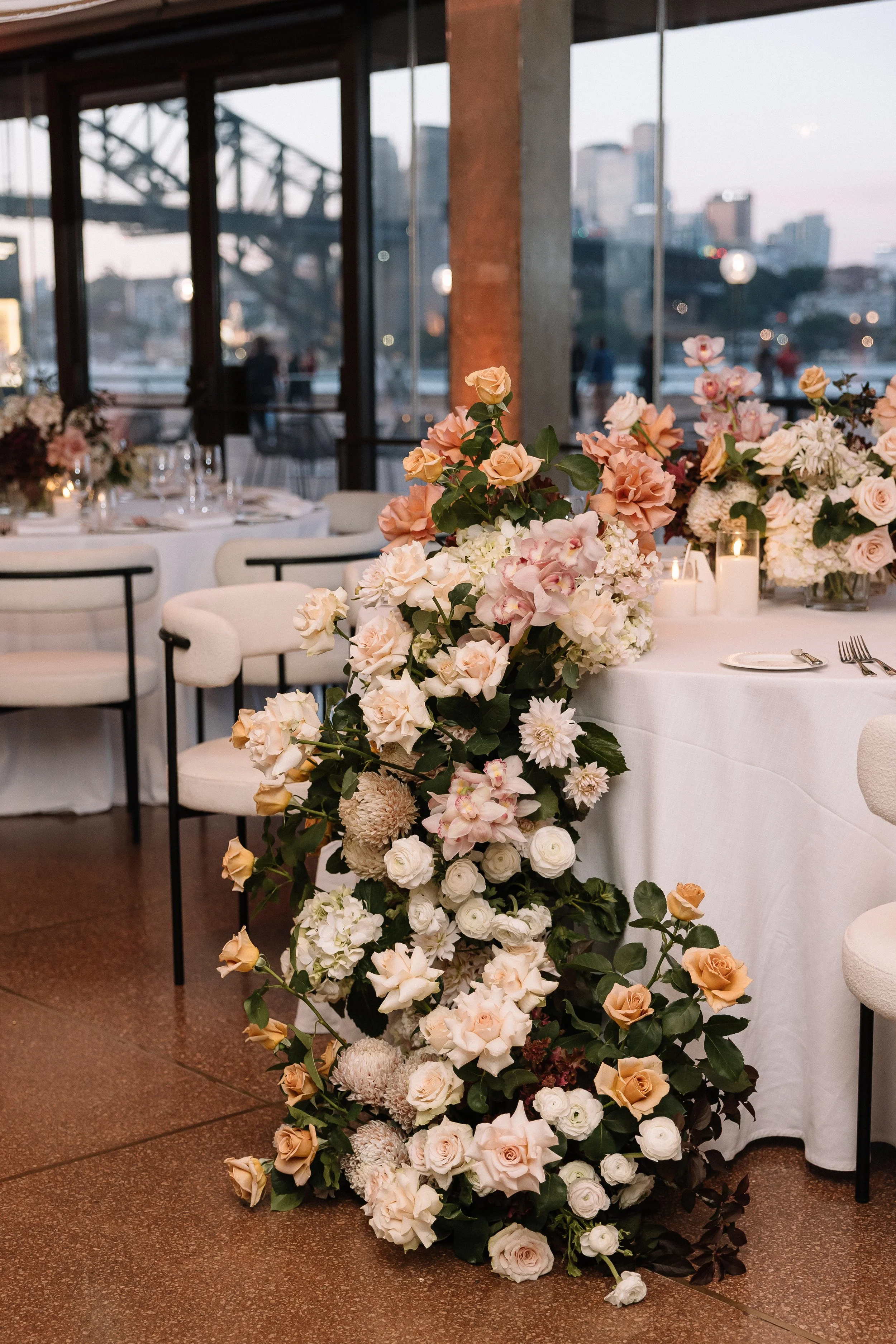 A floral arrangement of pastel-colored roses, peonies, and dahlias cascades over a white tablecloth at a wedding reception or event held in a glass-walled venue with city and bridge view.