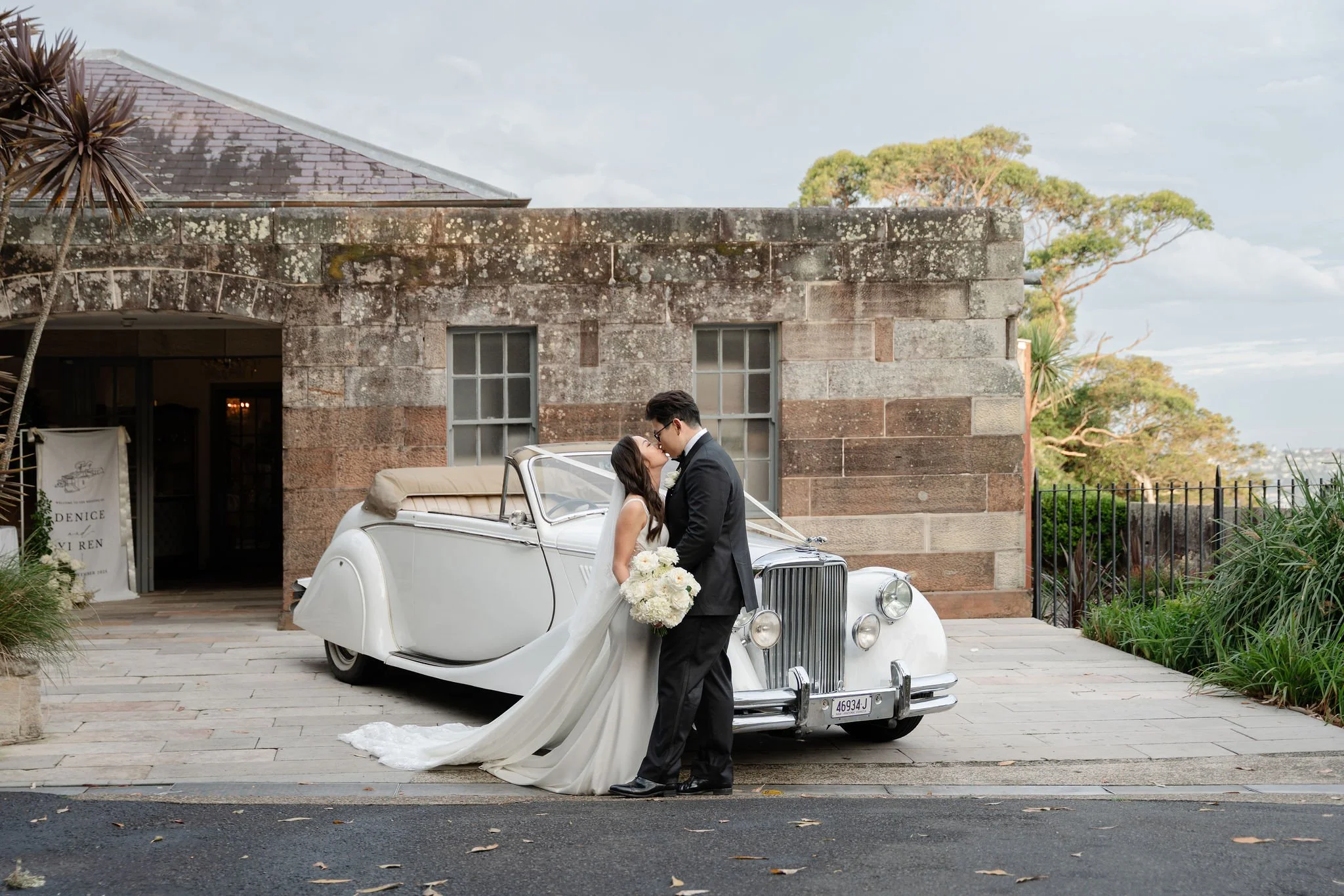 A bride and groom kissing next to a vintage white convertible car outside a brick building with trees in the background.