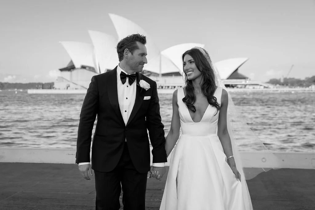 Black and white photo of a bride and groom holding hands and smiling at each other near a water body, with the Sydney Opera House in the background.
