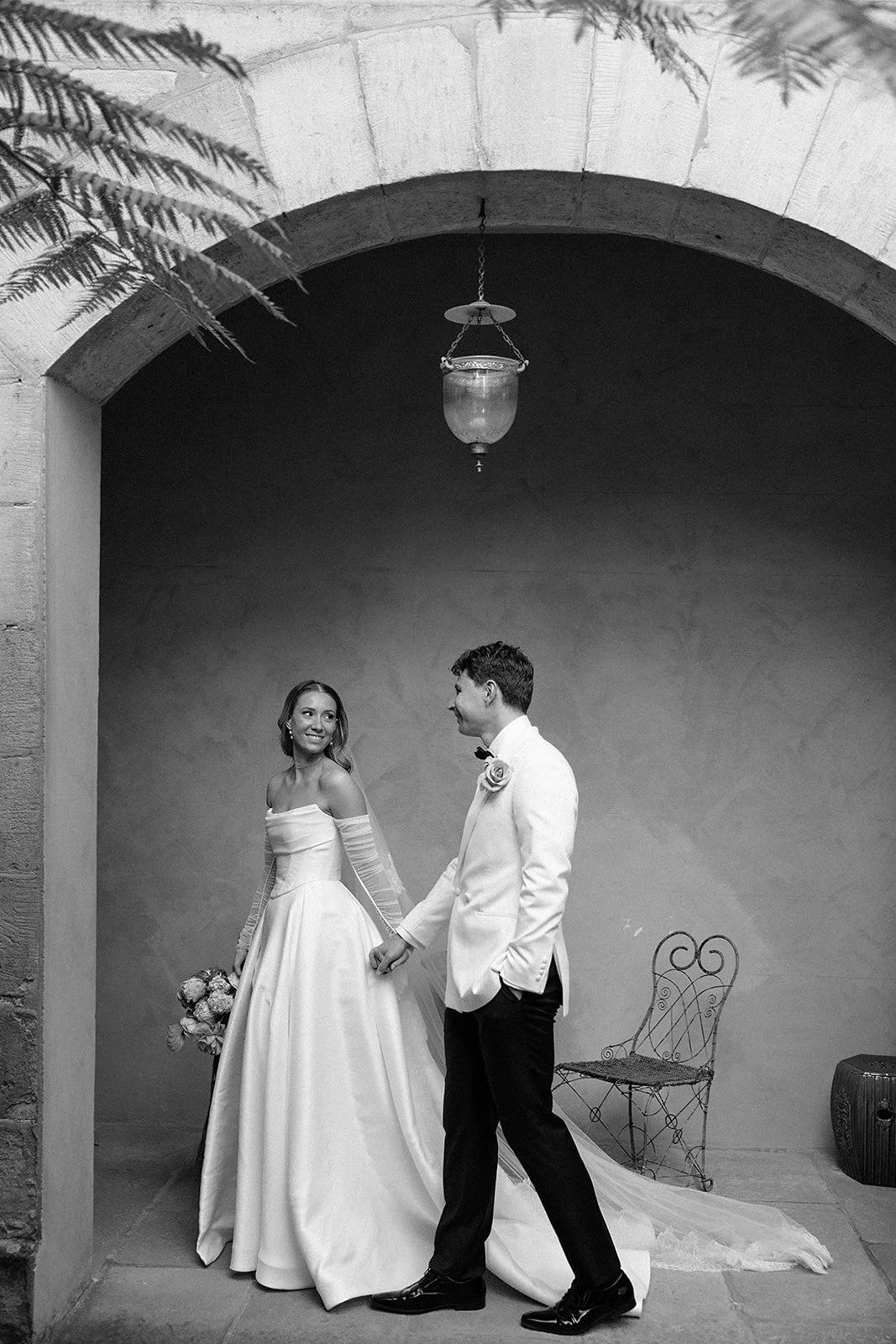 Black and white photo of a bride and groom holding hands, standing under a stone archway. The bride is wearing a long white wedding dress and holding a bouquet, smiling at the groom. The groom is dressed in a white tuxedo jacket with black pants, looking at the bride. There is a vintage chair and a textured wall in the background, with a hanging lantern above them.