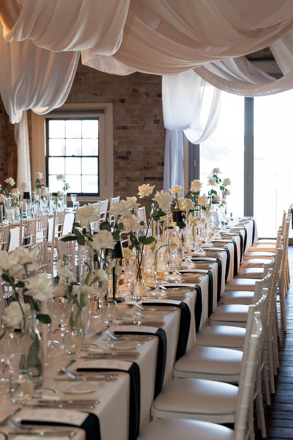 Elegant banquet table setup with white flowers, glassware, and silverware in a rustic venue with exposed brick walls and large windows with draped white curtains.