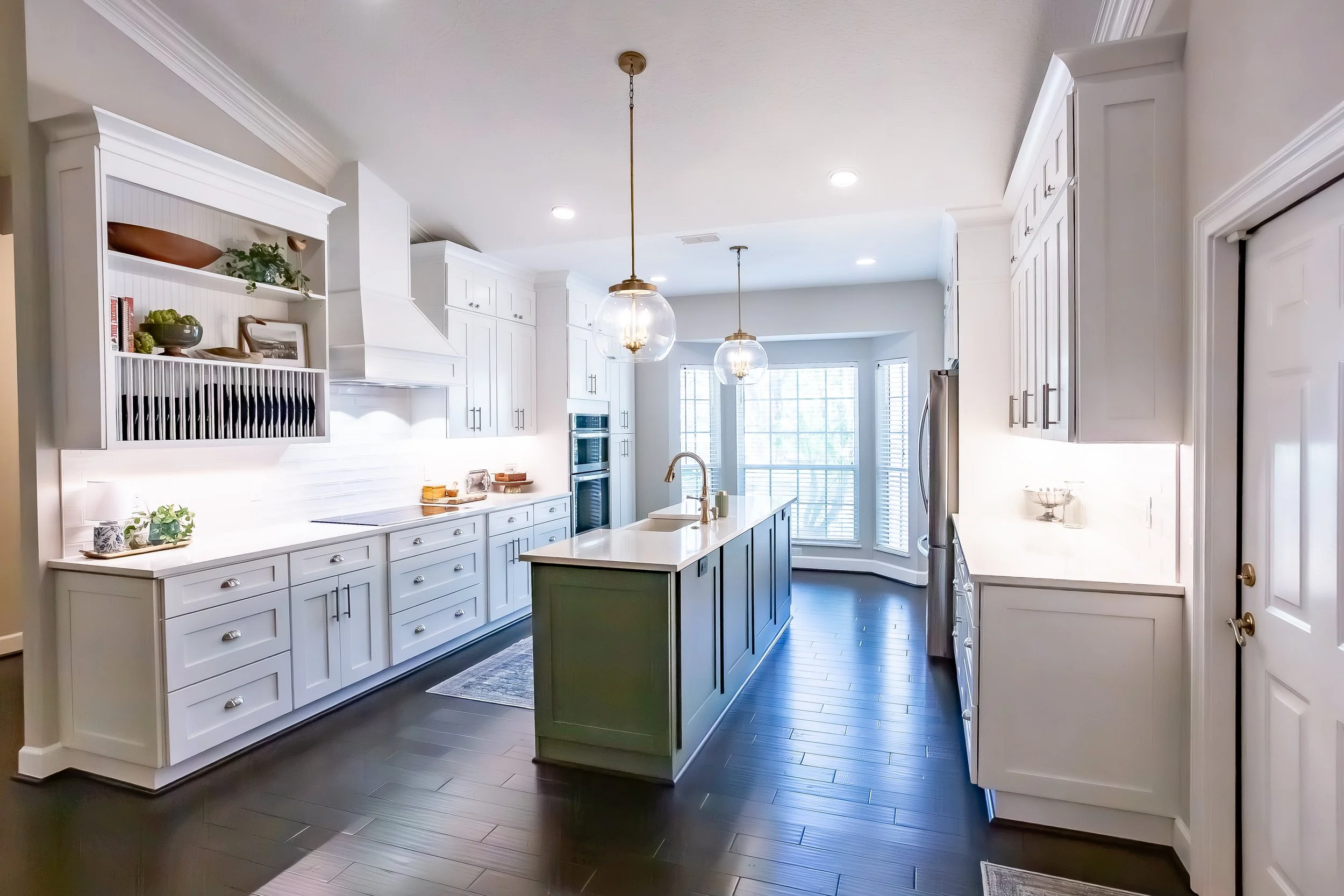 Bright and modern kitchen with white cabinets, a green island with a white countertop, pendant lighting, stainless steel appliances, and dark hardwood floors. Large bay windows in the background allow natural light to fill the space.
