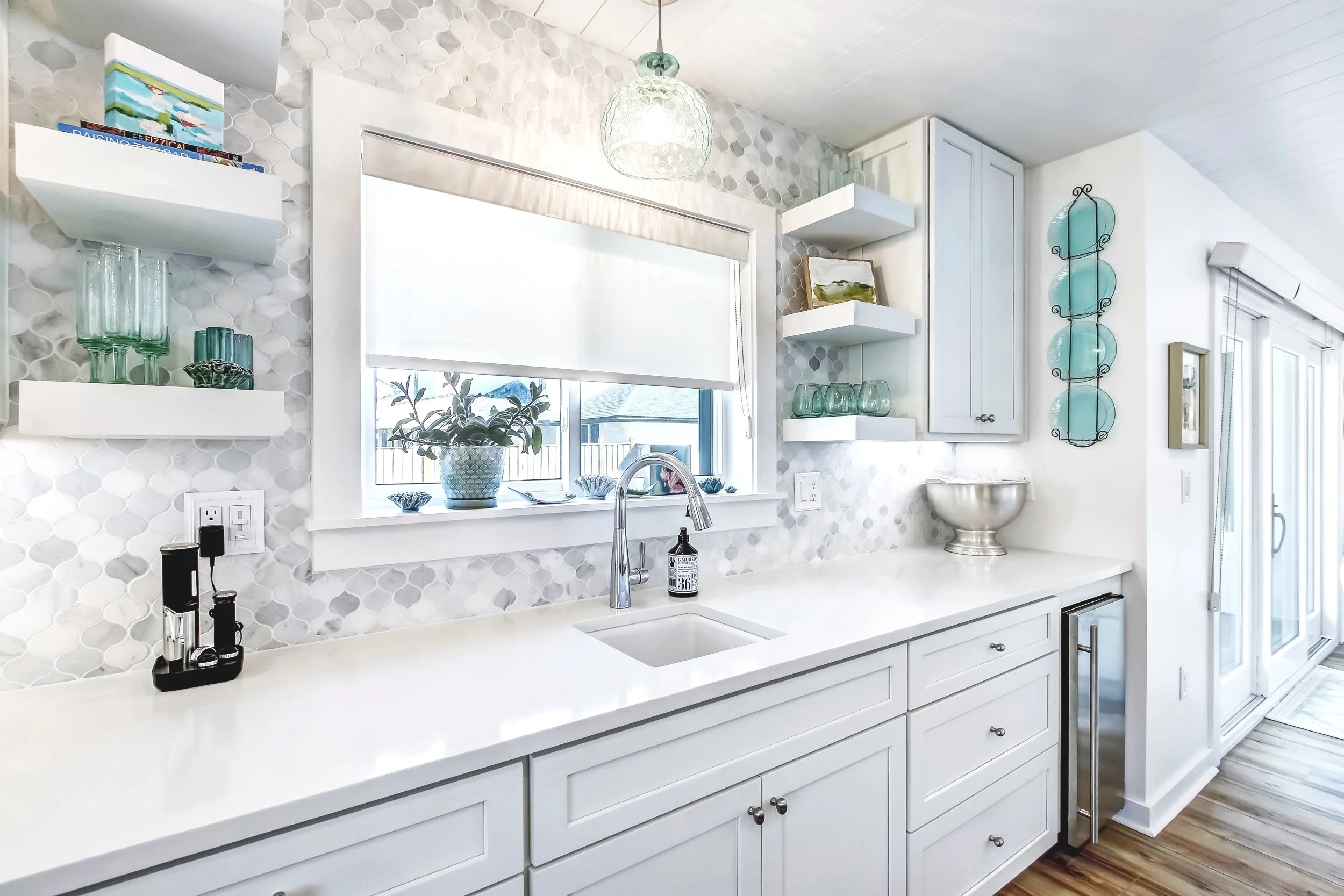 Modern kitchen with white cabinets, marble countertop, gray hexagonal tile backsplash, and a large window with a blind. Decor includes glassware, books, and plants. A stainless steel faucet and small appliances are on the counter.