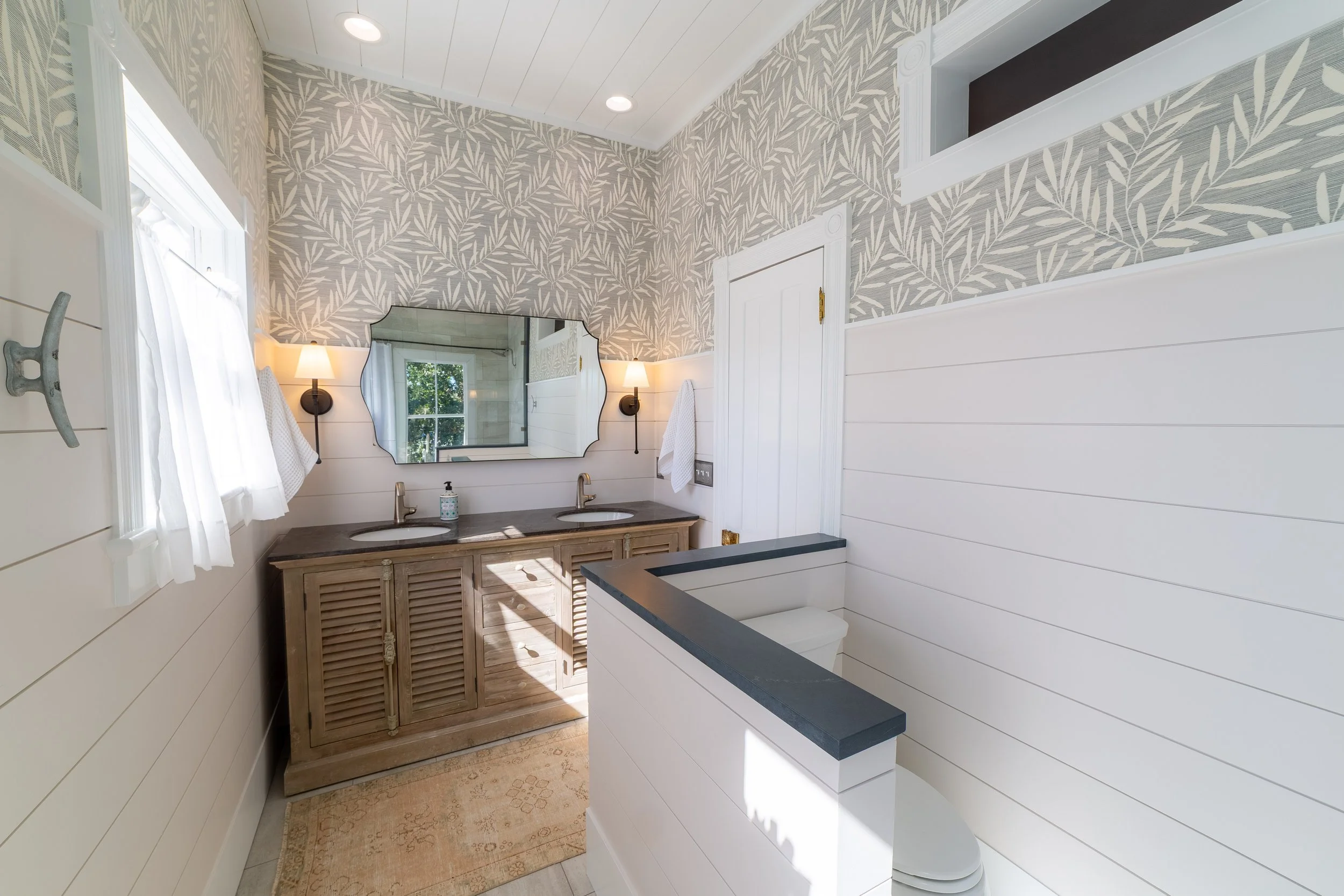 Coastal bathroom with white shiplap walls, tropical leaf wallpaper, double vanity, and closed off toilet.