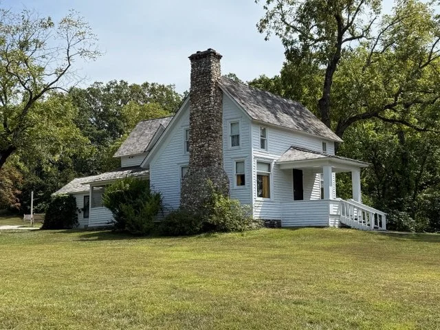 White two-story farmhouse with steep gabled roof and prominent stone chimney, featuring a covered front porch with white railings, surrounded by mature trees and a well-maintained lawn.