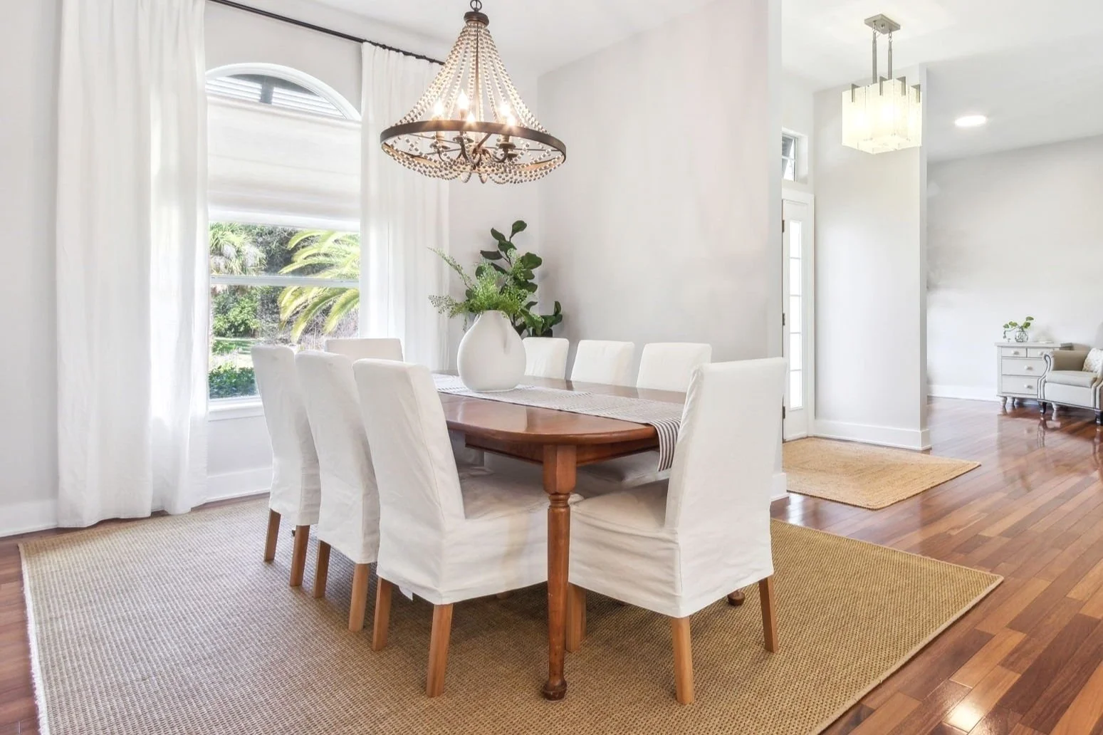 Bright dining room with a wooden table, six white slipcovered chairs, a chandelier, large windows with curtains, and a plant in a vase on the table.