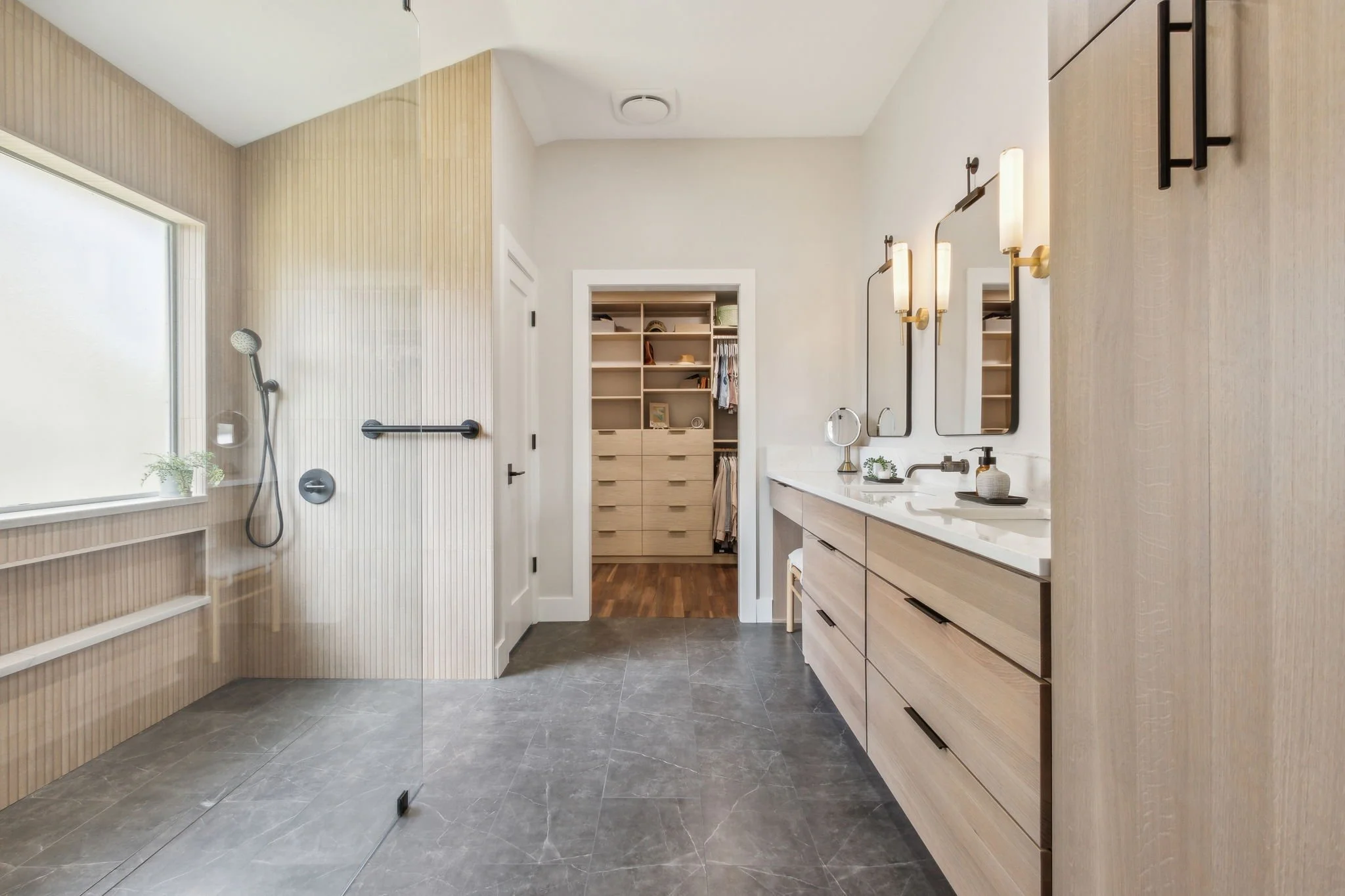 Modern bathroom with walk-in shower, floating vanity with dual sinks, and mirrors. Features wooden cabinetry and open door to walk-in closet. Large window and sleek pendant lights enhance the decor.