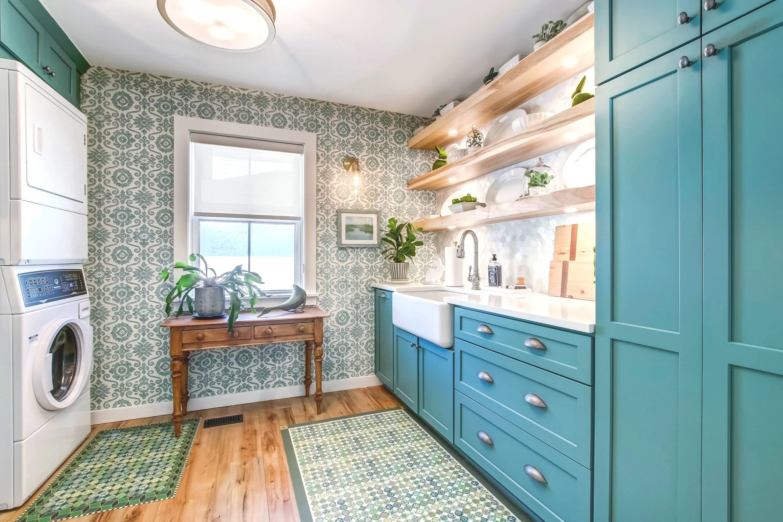Laundry room with teal cabinets, patterned wallpaper, a stacked washer and dryer, farmhouse sink, wooden shelves with dishes, and a wooden table with plants.