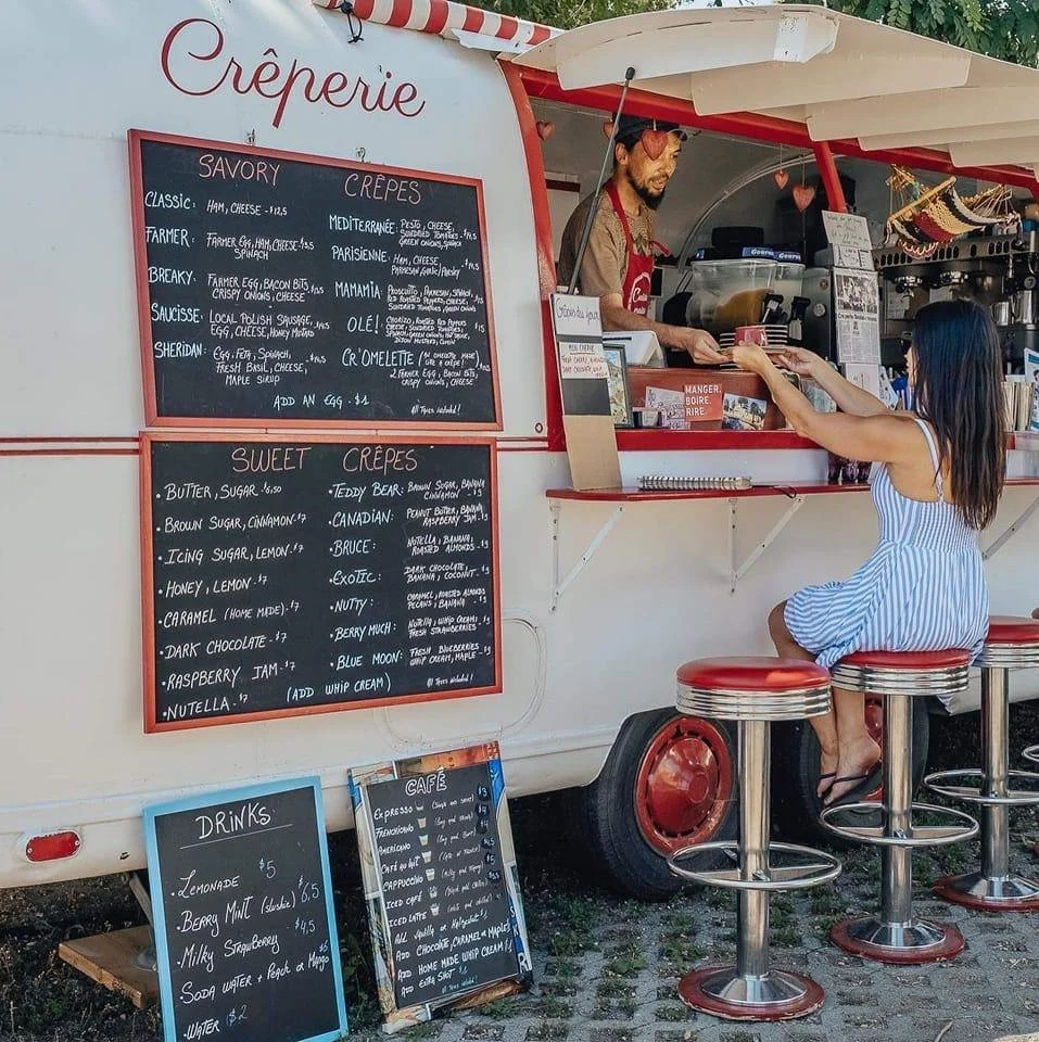 A woman in a blue and white striped dress ordering at a food truck creperie with a man inside taking her order. The menu boards list various sweet and savory crepes, drinks, and cafe options.