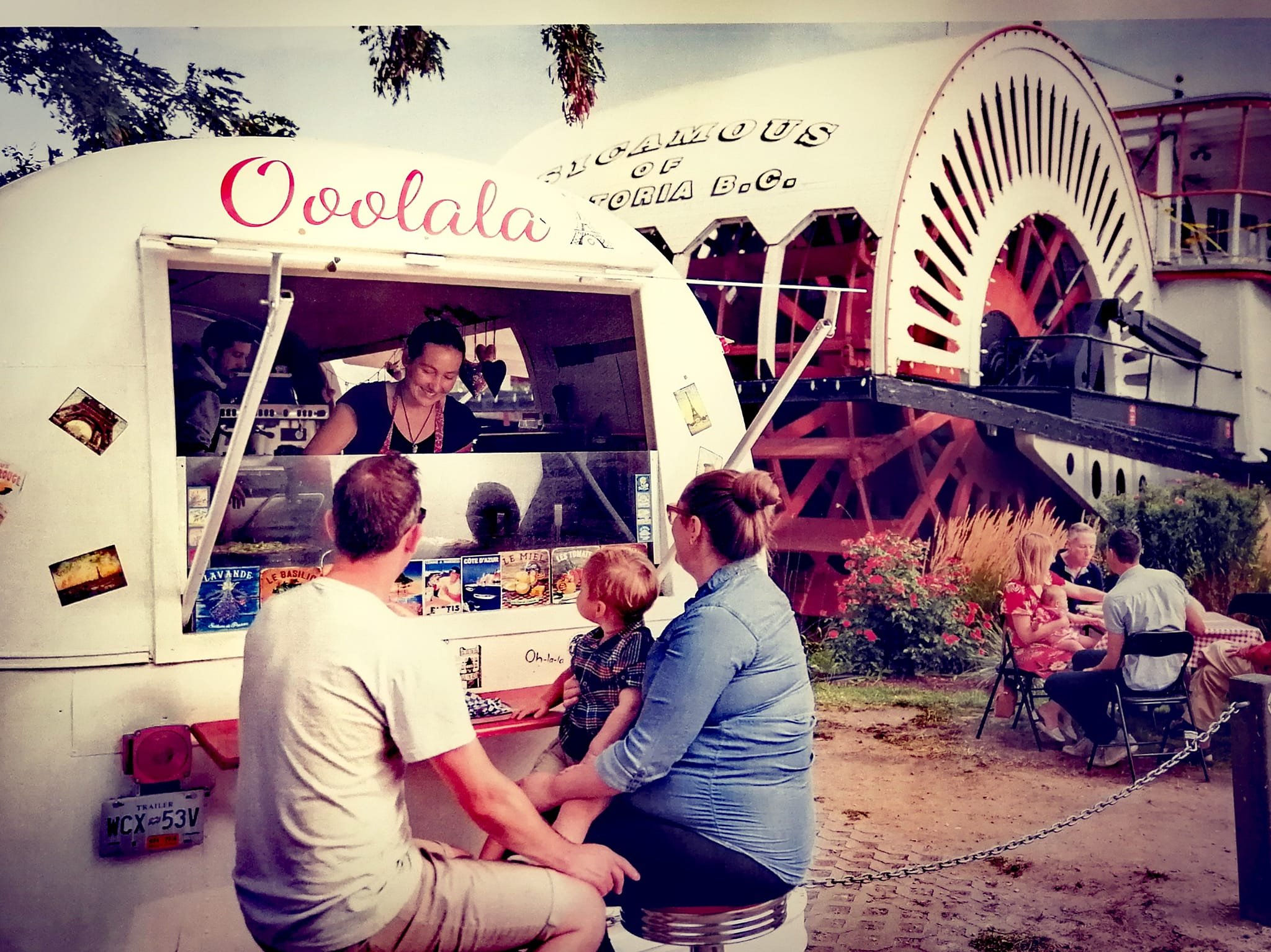 People sitting outside at a vintage drive-in food stand with a large white trailer. The stand has the word 'Oodaloo' on it, and there are various posters on the front. Customers are seated at tables with a woman taking orders inside the stand.