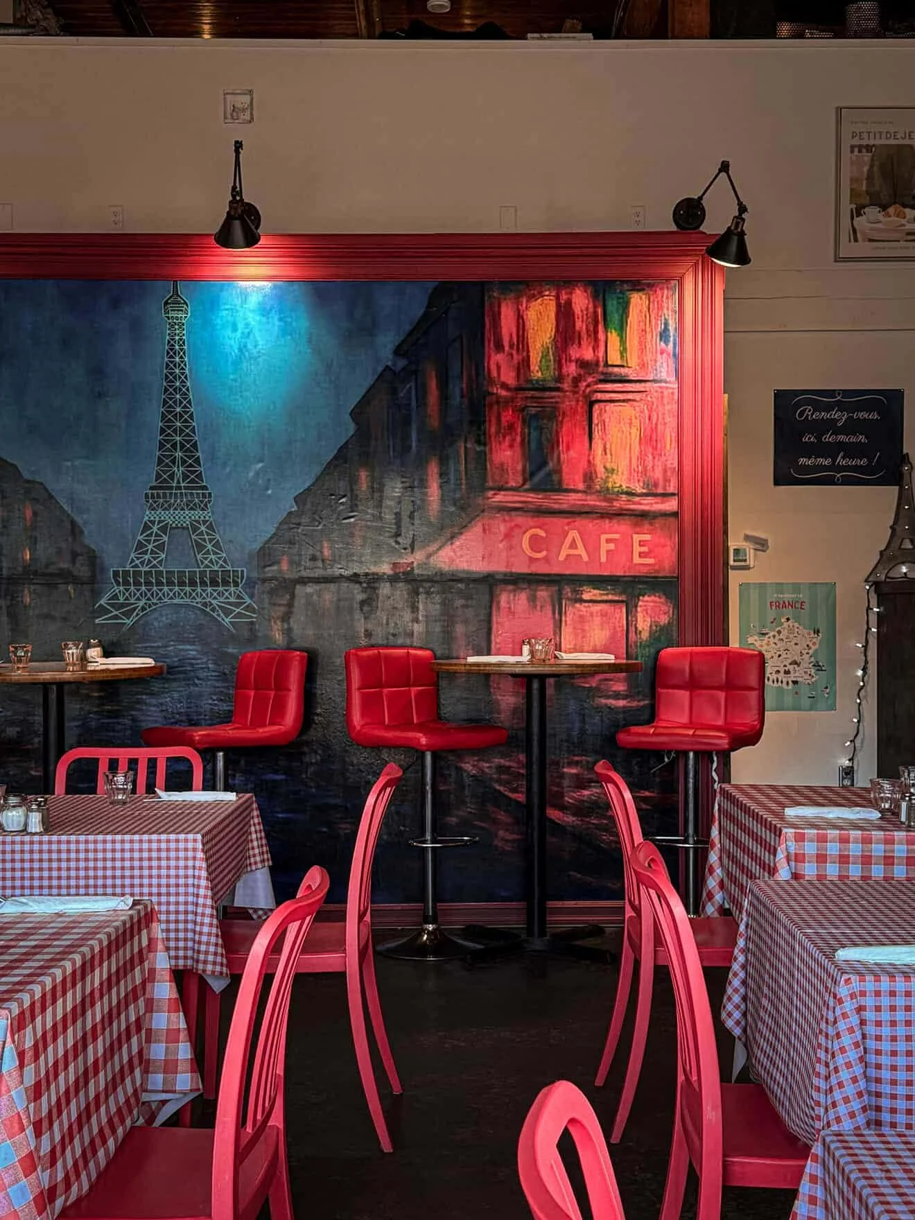 Interior of a French-themed cafe with red chairs, checkered tablecloths, and a large mural of the Eiffel Tower and Paris streets on the wall.