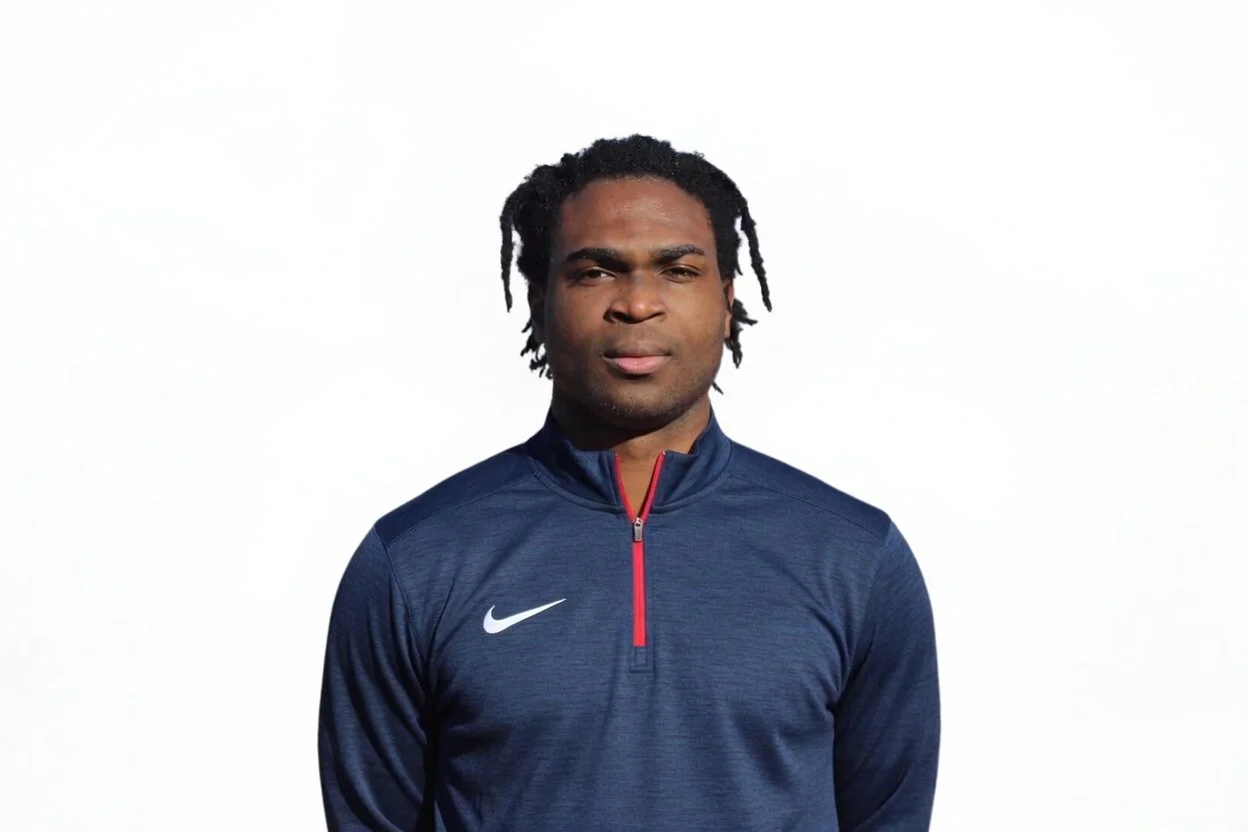A man with braided hair wearing a navy blue jacket, smiling.
