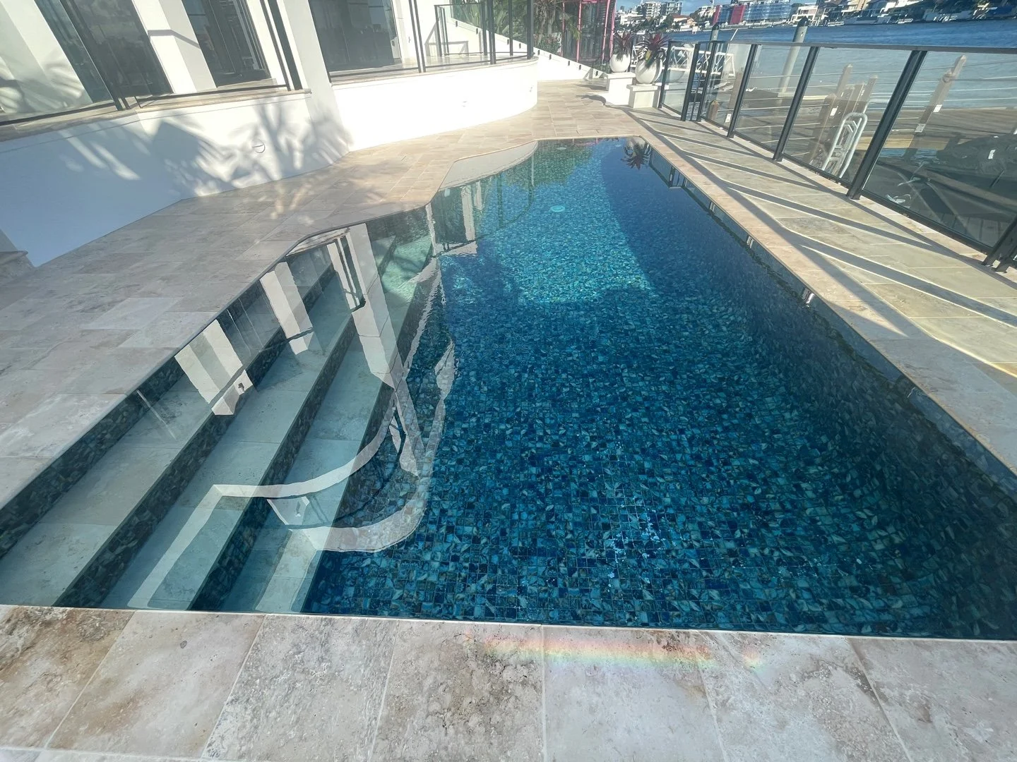 A rooftop swimming pool with blue mosaic tiles, surrounded by a stone deck and glass railings, overlooking an ocean view.