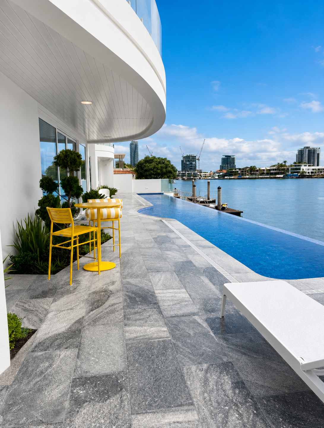 Modern residential balcony with yellow outdoor furniture, potted plants, an infinity pool, and view of city skyline and waterway against blue sky.
