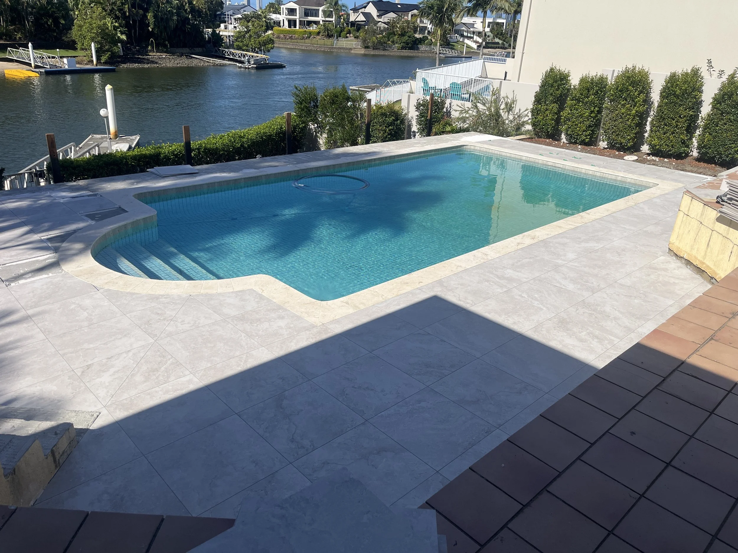 A backyard pool area with a swimming pool surrounded by tiled decking. In the background, there is a waterway with boats docked along the shore and residential buildings with lush greenery and palm trees.