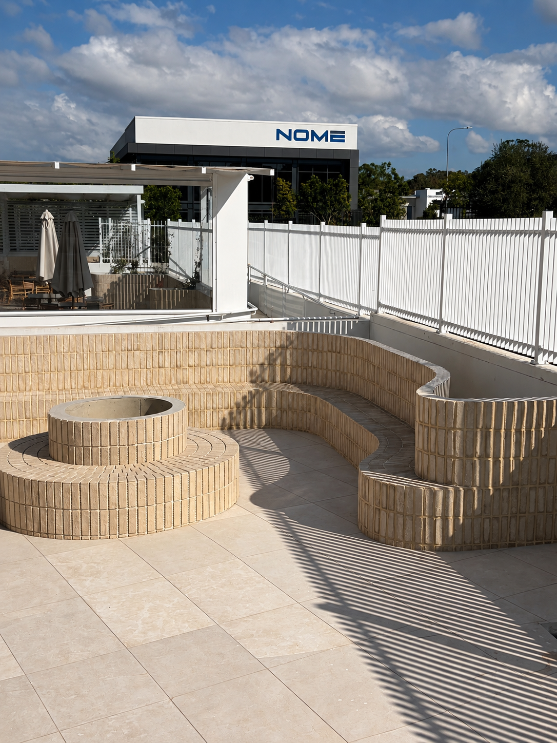 Empty outdoor pool area with beige brick surround, white fence, and adjacent patio with table, umbrellas, and chairs. Modern building with sign reading 'NOME' in the background, under partly cloudy sky.