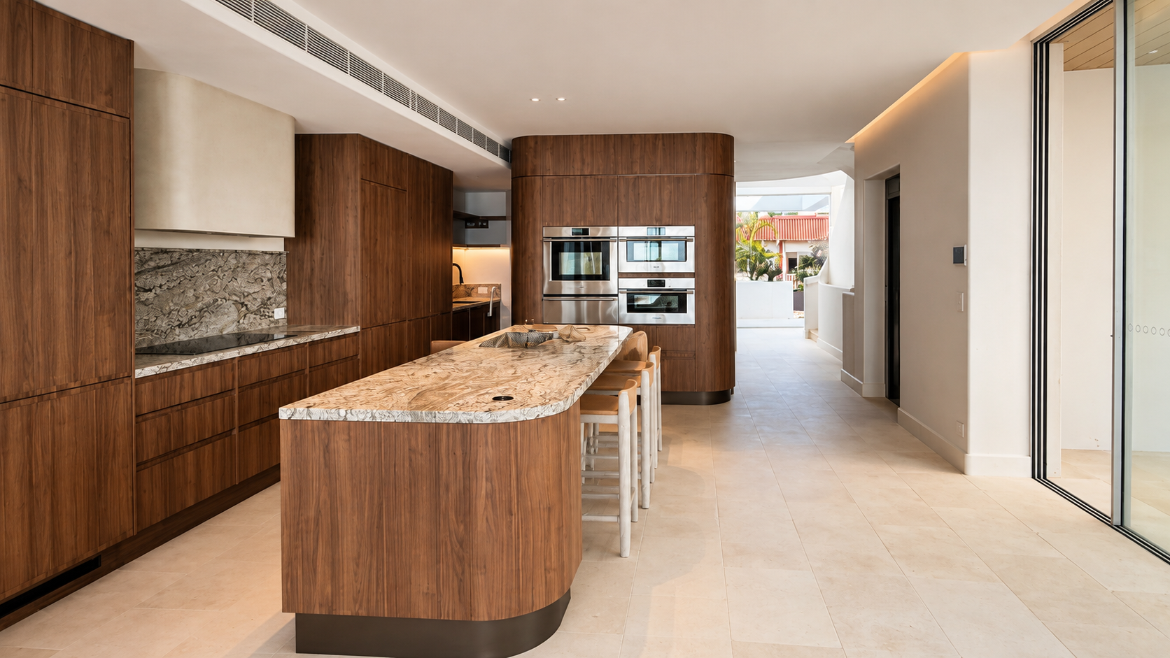 Modern kitchen with wooden cabinetry, a large marble island, built-in stainless steel appliances, and a view outside through glass doors.