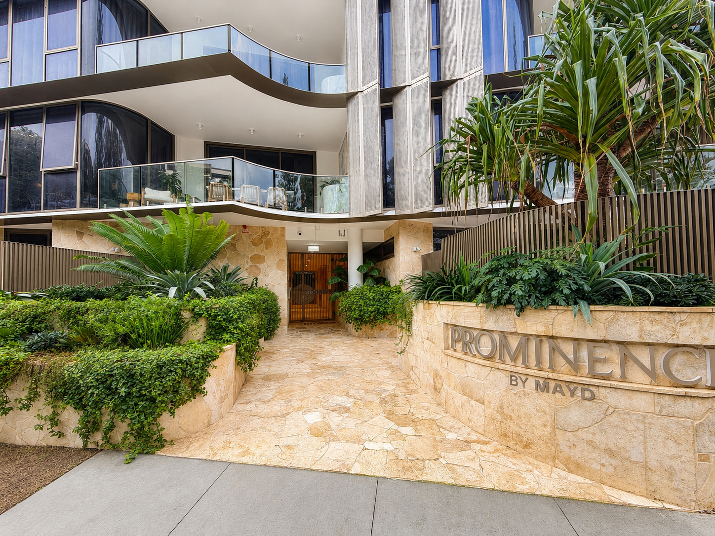 Entrance to the Prominence by MAYD residential building with lush greenery and a stone pathway leading to the door.