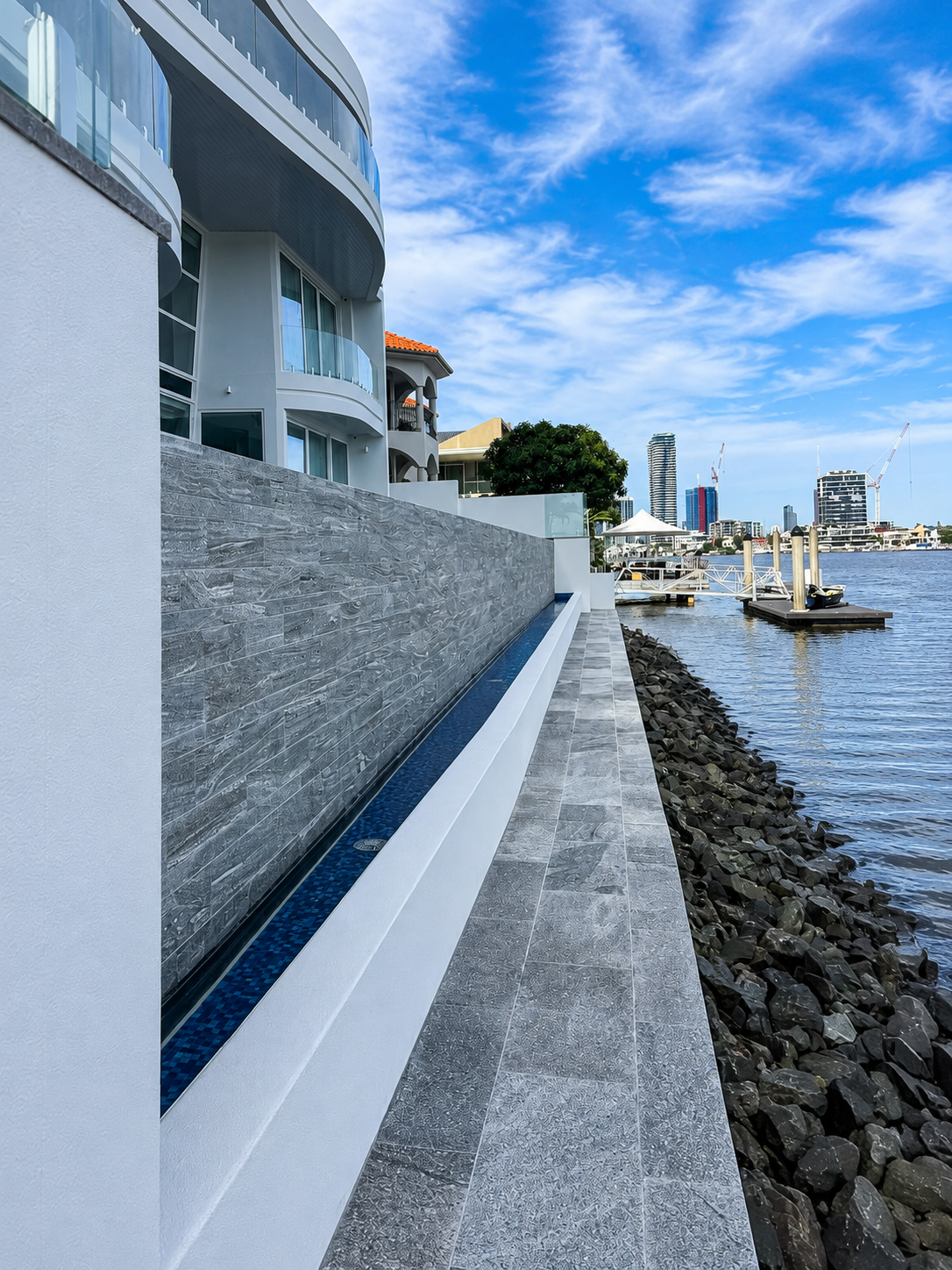 Modern waterfront building with glass balconies, gray tiled walkway, and a rocky shoreline beside a river with city skyline in the background.